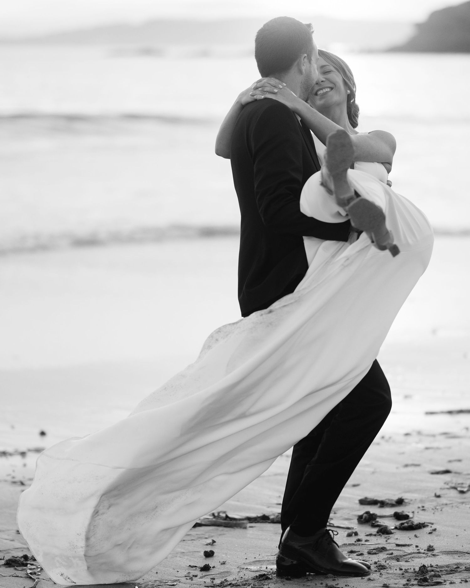 A joyful black and white beach wedding photo capturing a playful lift moment with flowing white dress.