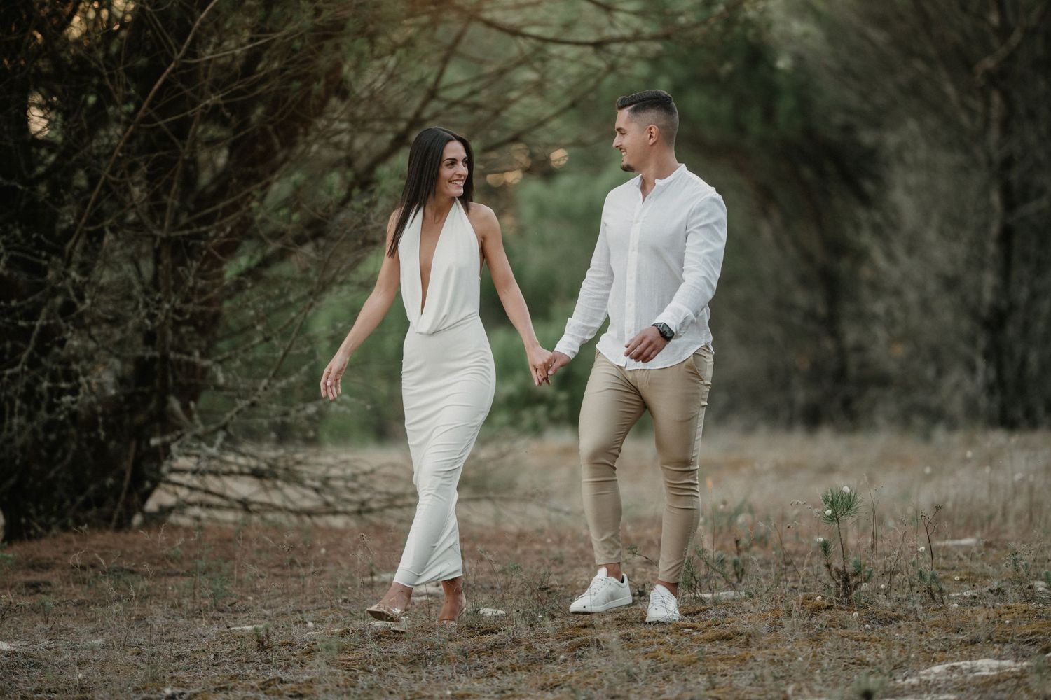 A couple in white outfits walks together through a dreamy forest setting during a romantic photoshoot.