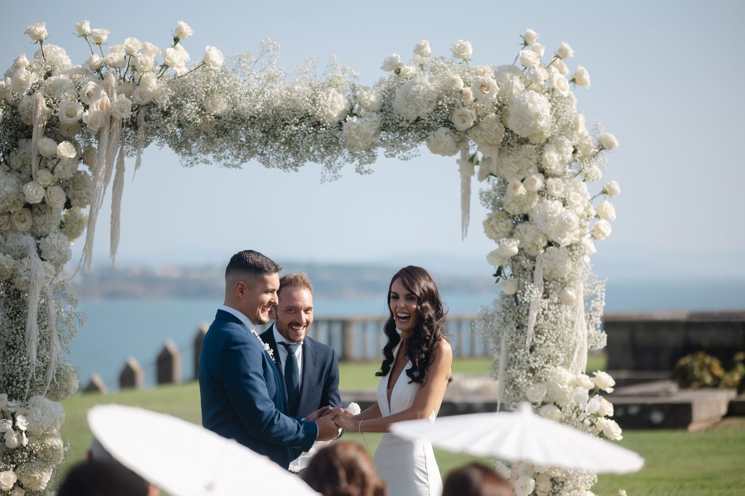 Wedding ceremony takes place under an elegant white floral arch overlooking a scenic waterfront view.