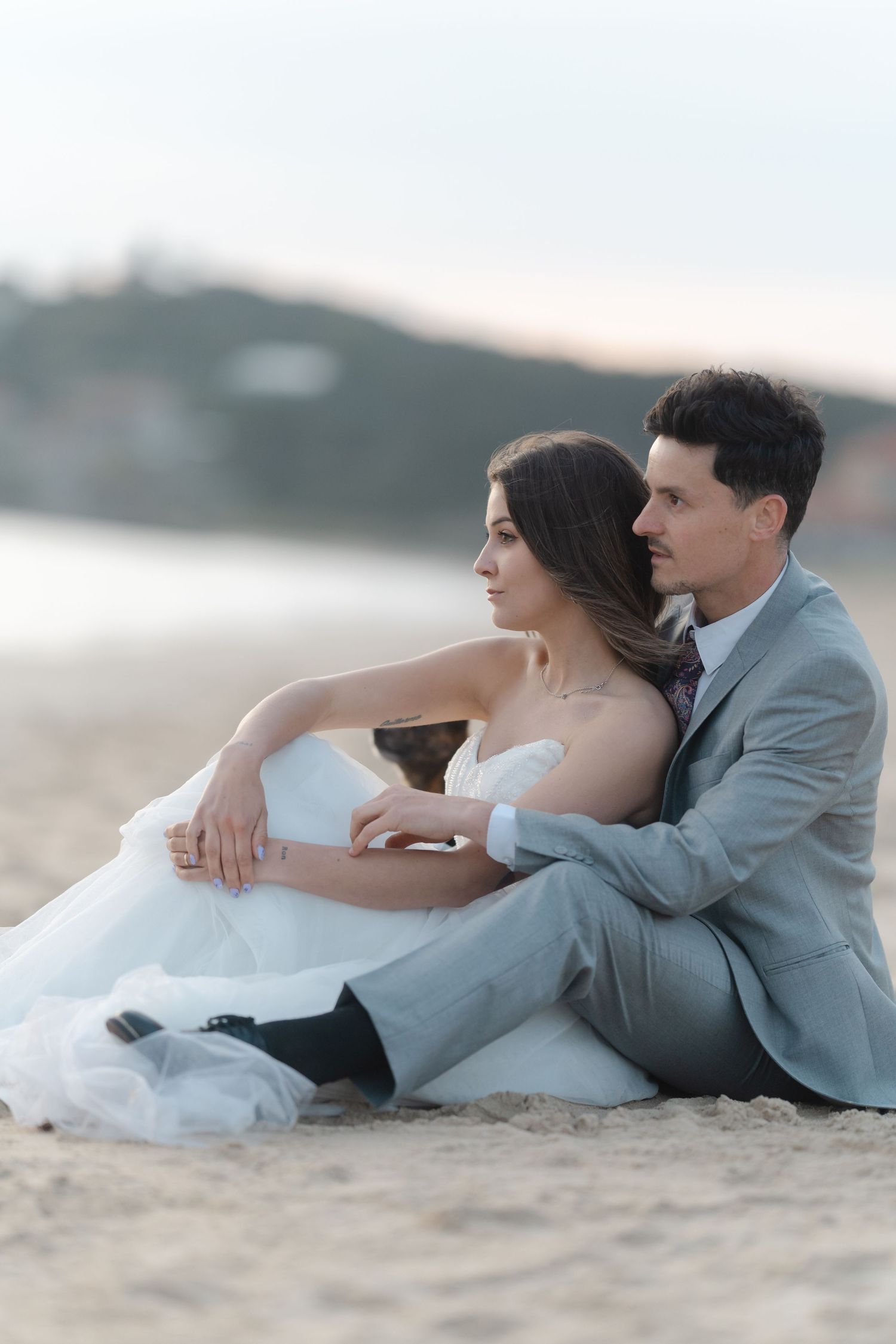 Bride and groom sitting together on sandy beach in wedding attire, gazing into the distance at sunset.