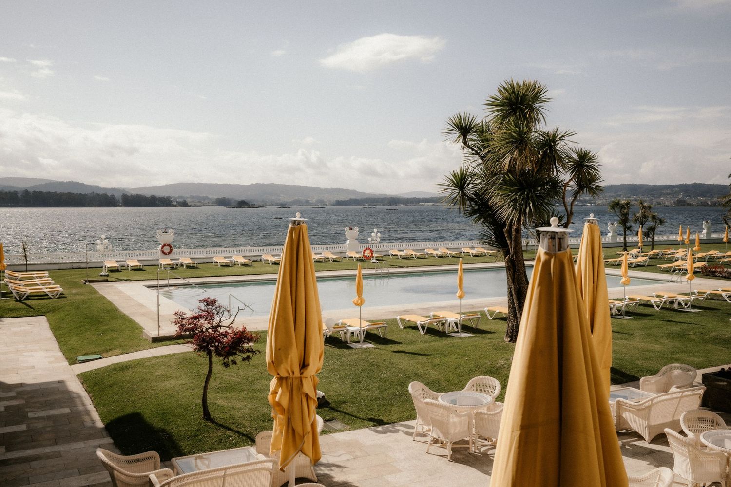 Panoramic waterfront restaurant terrace with yellow umbrellas overlooking a bay and mountains in the distance.