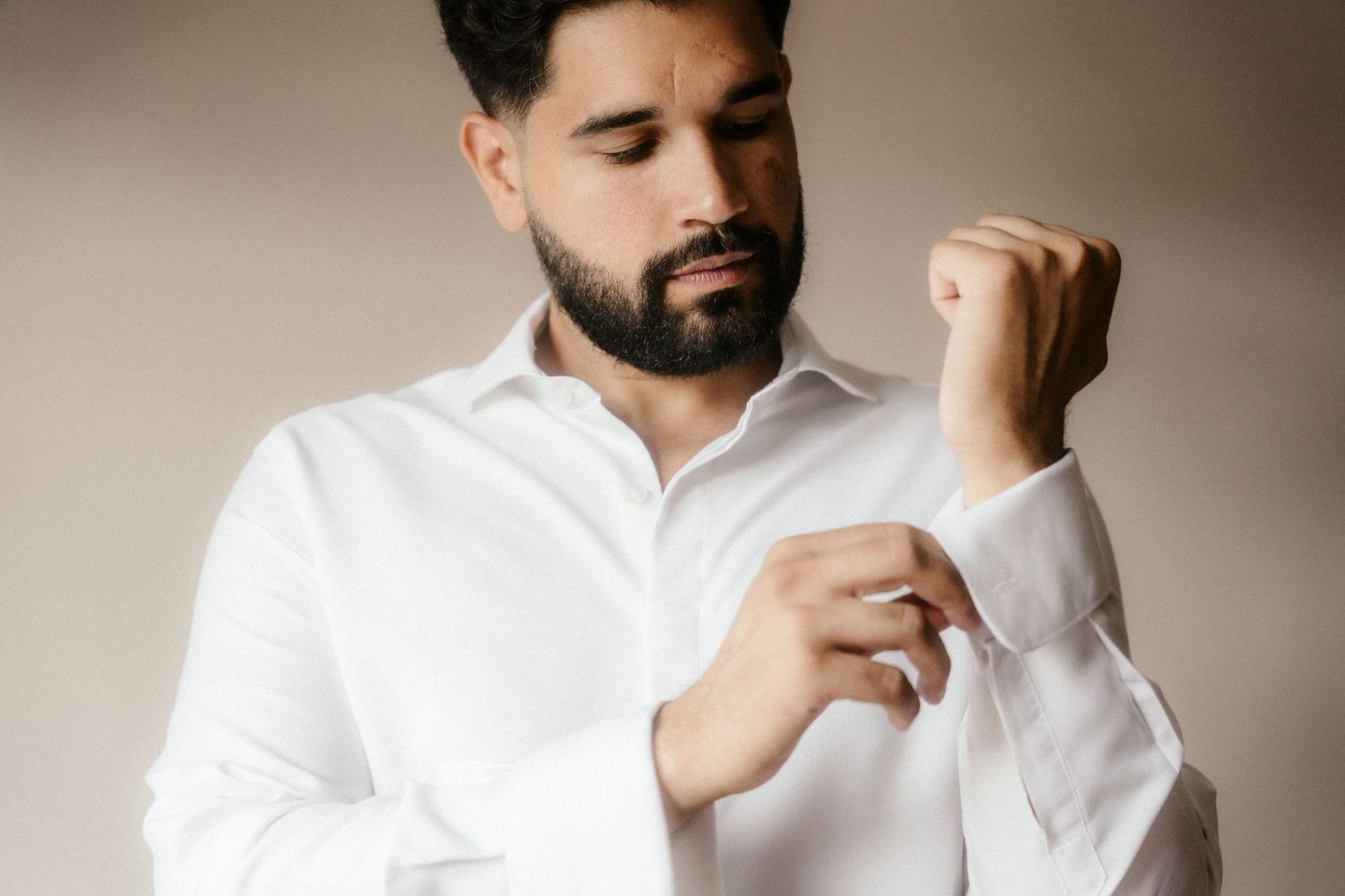 A man in a crisp white button-down shirt adjusts his collar while looking away from the camera in a studio setting.