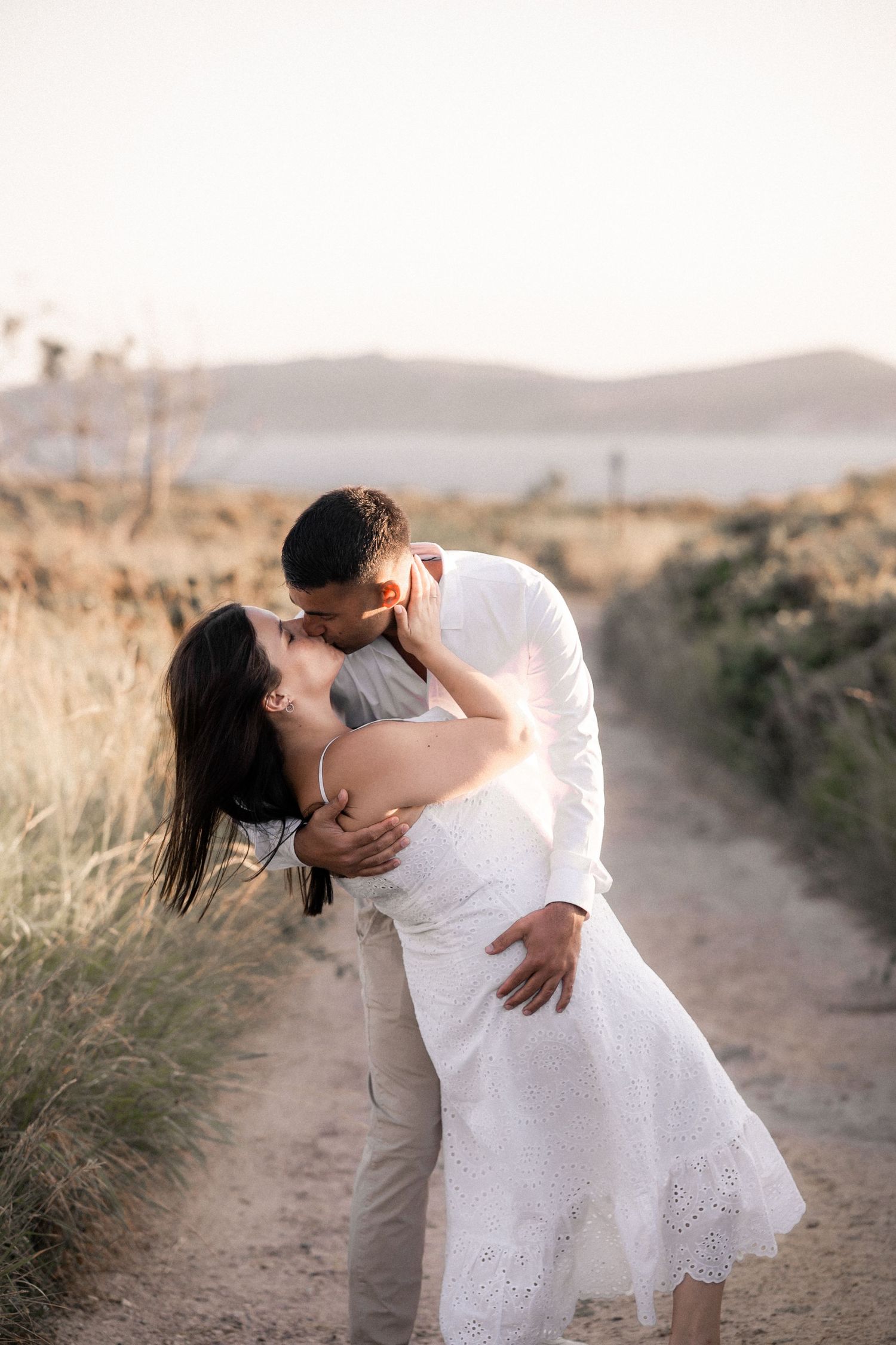 A romantic couple embraces on a dirt path surrounded by tall grass during golden hour.