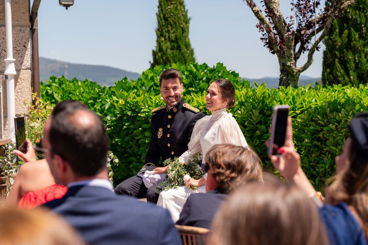 Wedding ceremony guests seated outdoors in a scenic garden venue with cypress trees and mountains in background.
