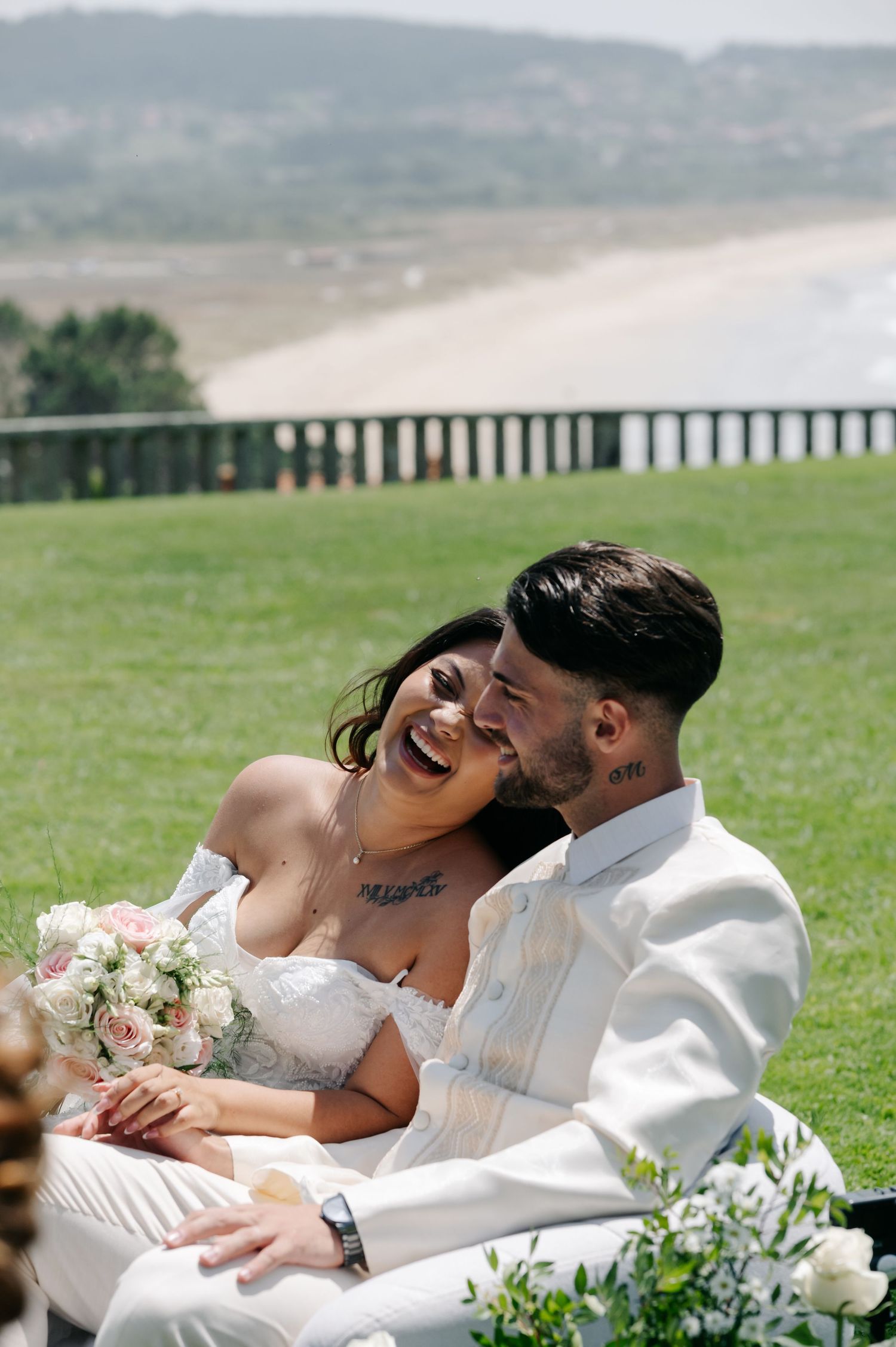 A couple shares a tender moment laying together in the grass with a bouquet of pink and white flowers nearby.