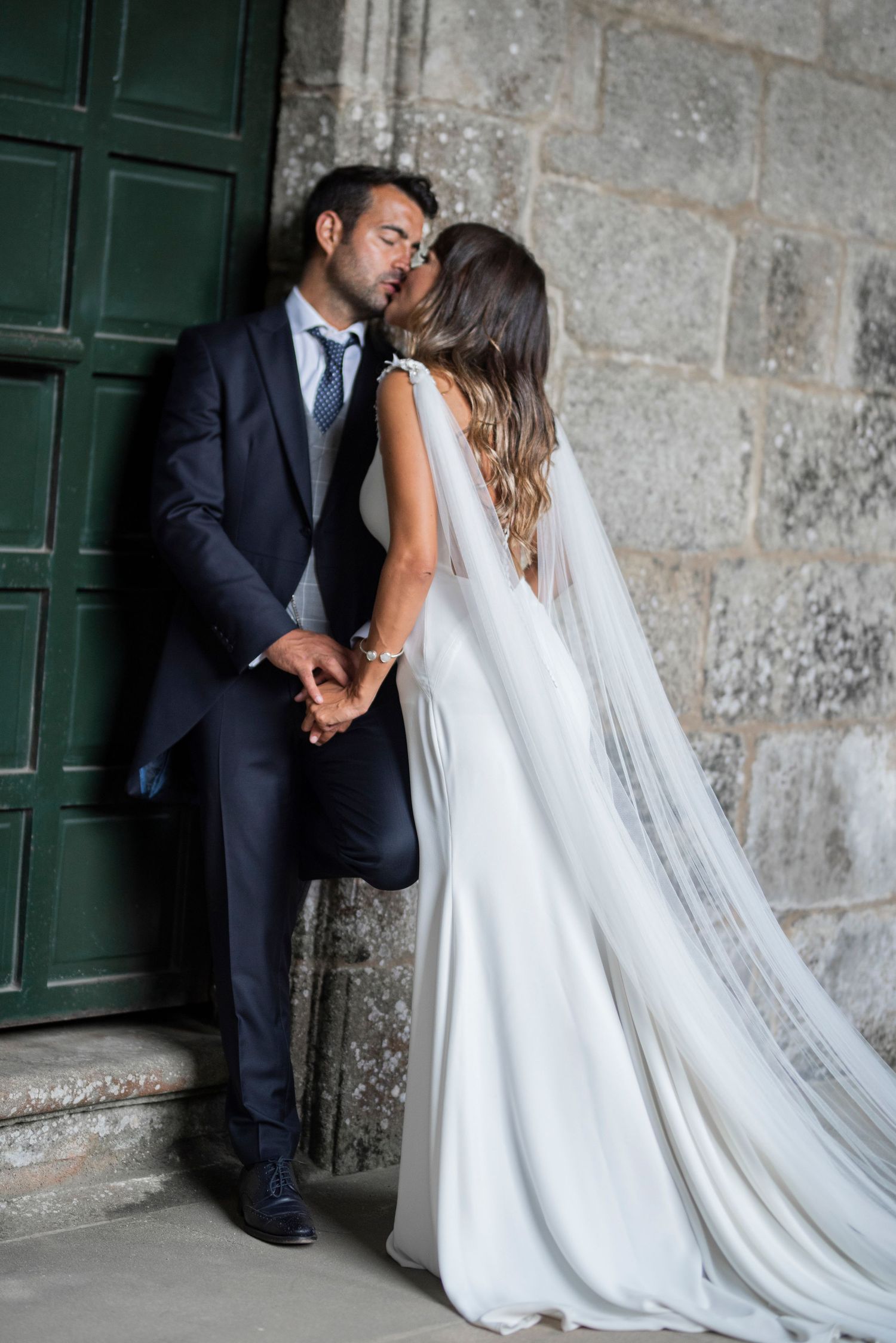 A romantic wedding couple shares intimate moments against a rustic stone wall with green doors.