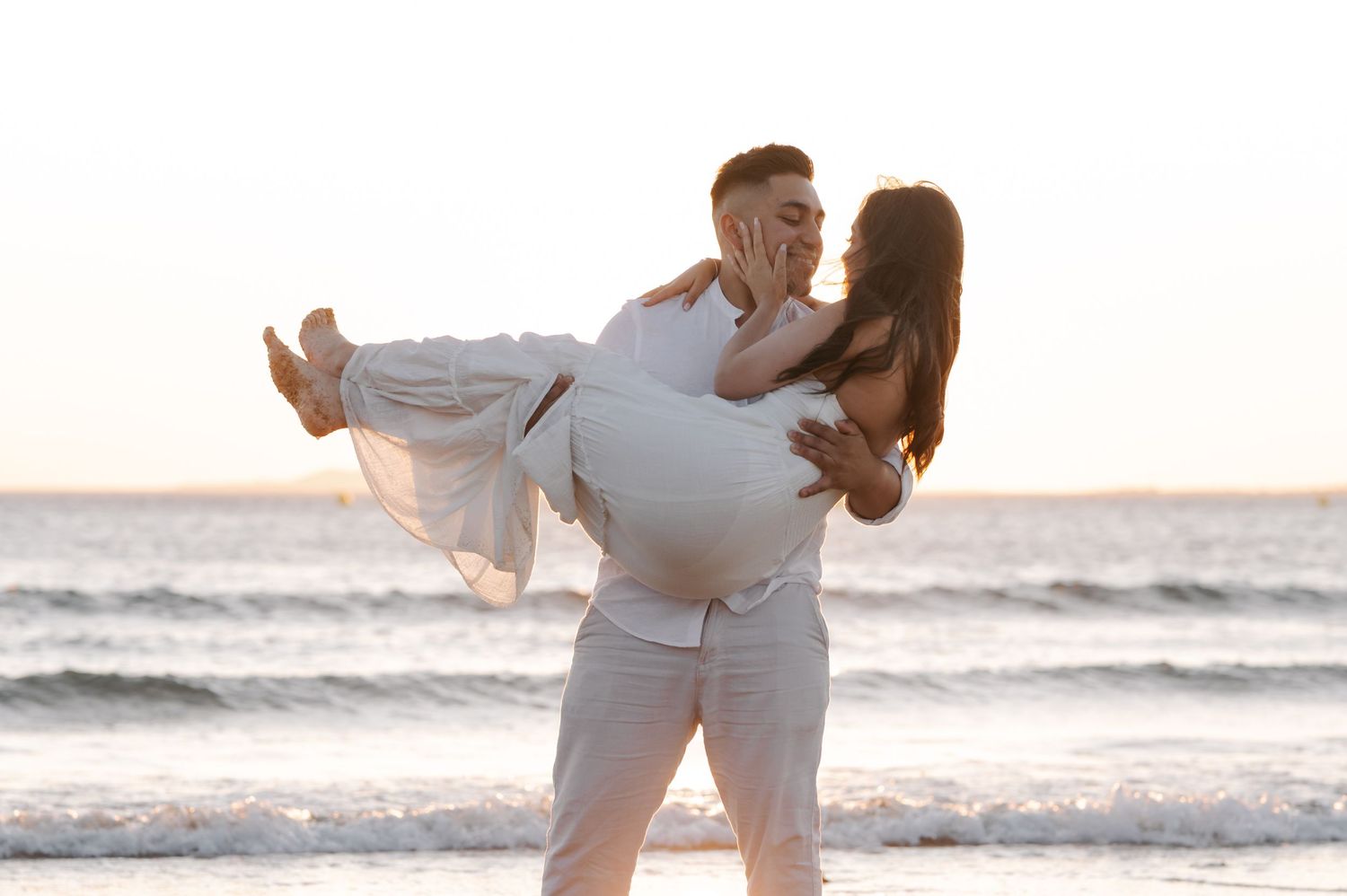 Couple in white outfits dances playfully on a sandy beach at sunset with ocean waves in the background.