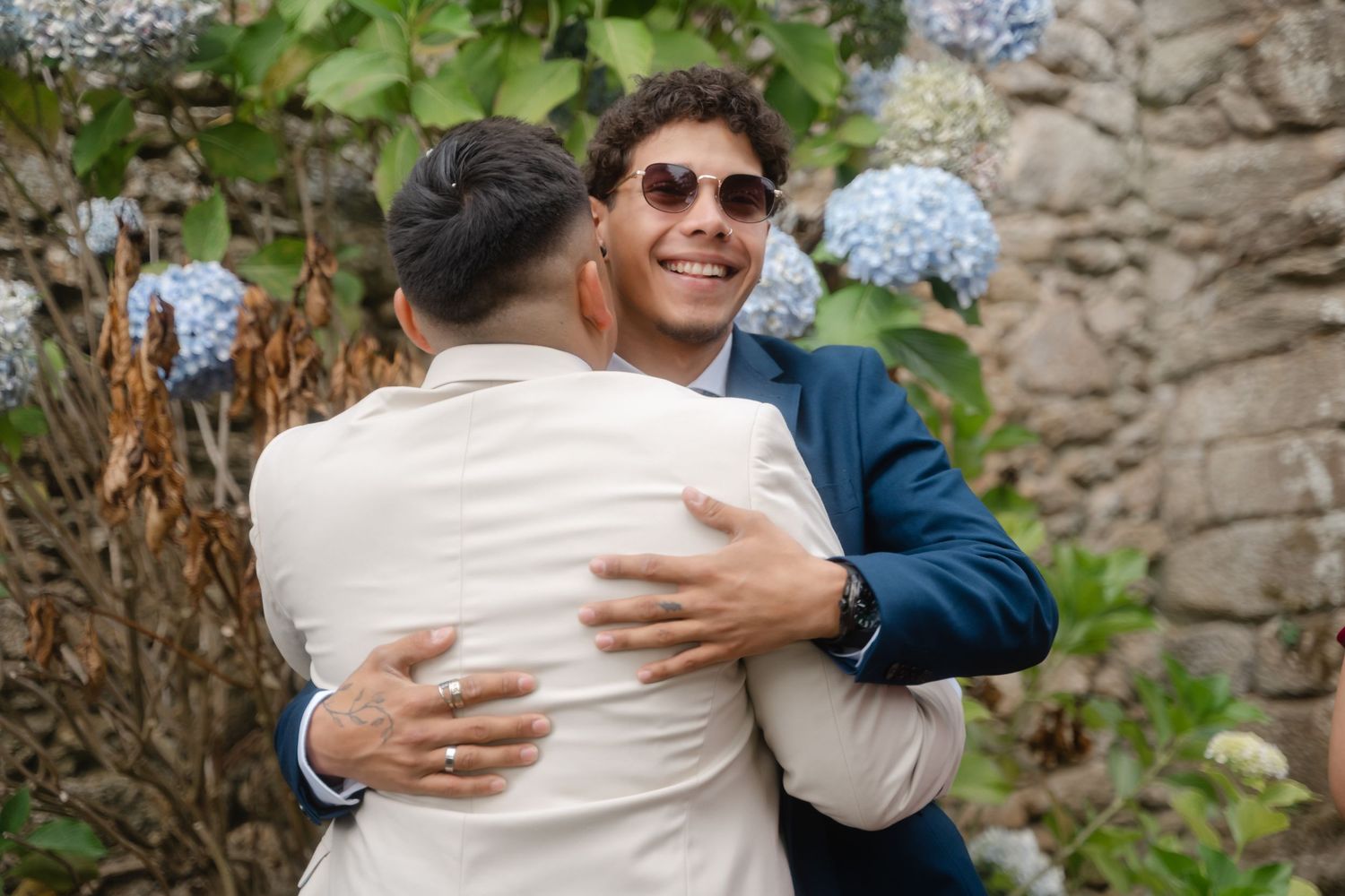 Wedding guests share warm embraces during outdoor celebration near stone wall.