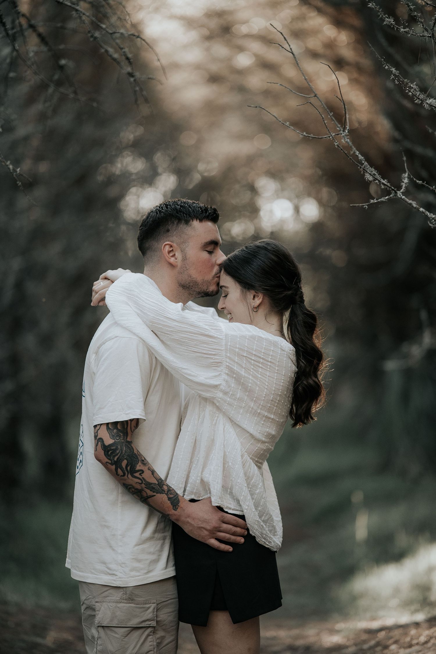 Romantic couple in white sweaters embrace tenderly in a moody forest setting during an intimate photo session.