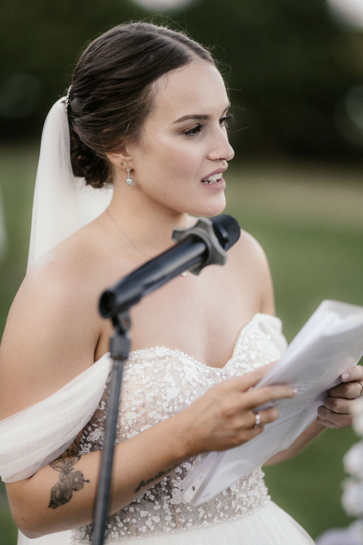 A bride in a beaded strapless gown reads from papers at an outdoor wedding ceremony.