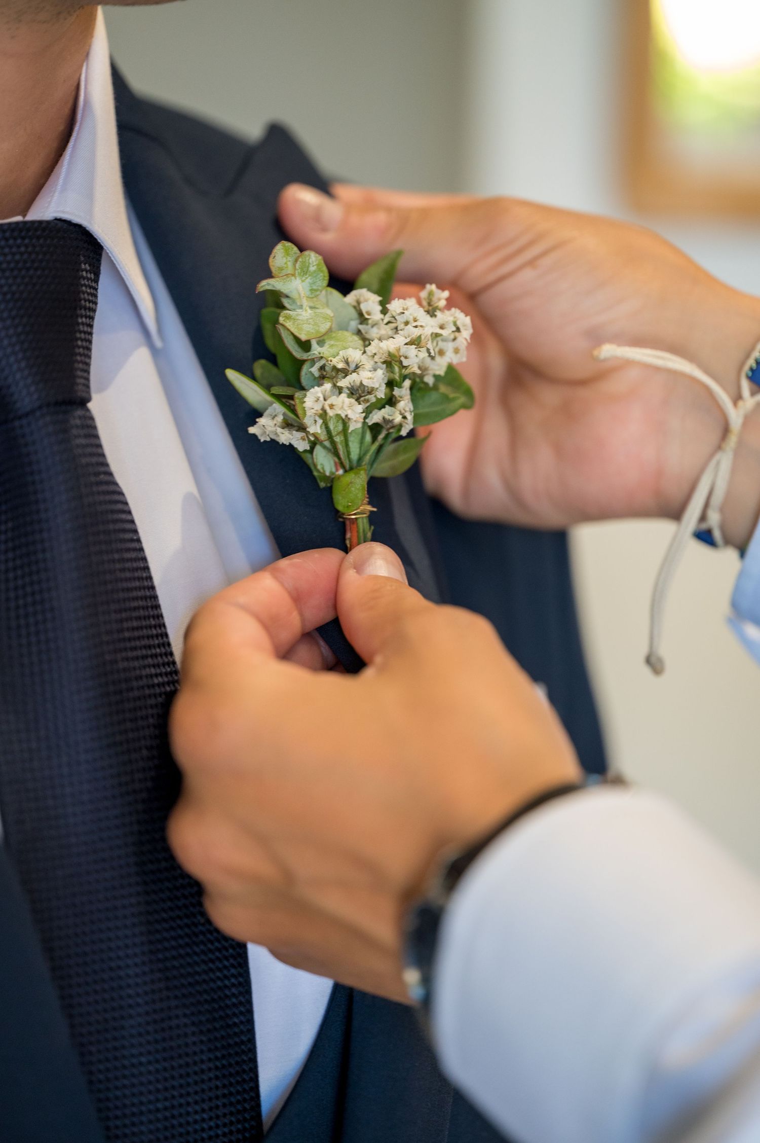 Hands pinning a delicate white and green boutonniere onto a navy suit lapel.