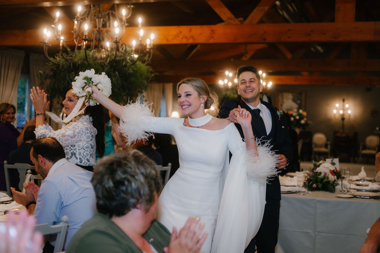 A romantic candlelit wedding reception with guests celebrating at tables in a rustic wooden barn venue.