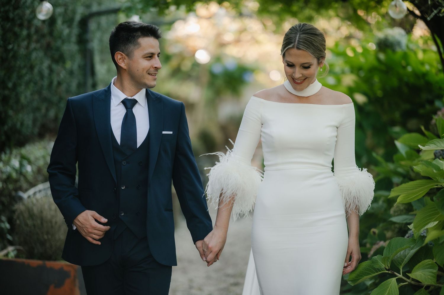 A couple in wedding attire walks hand in hand through a lush garden setting during their special day.