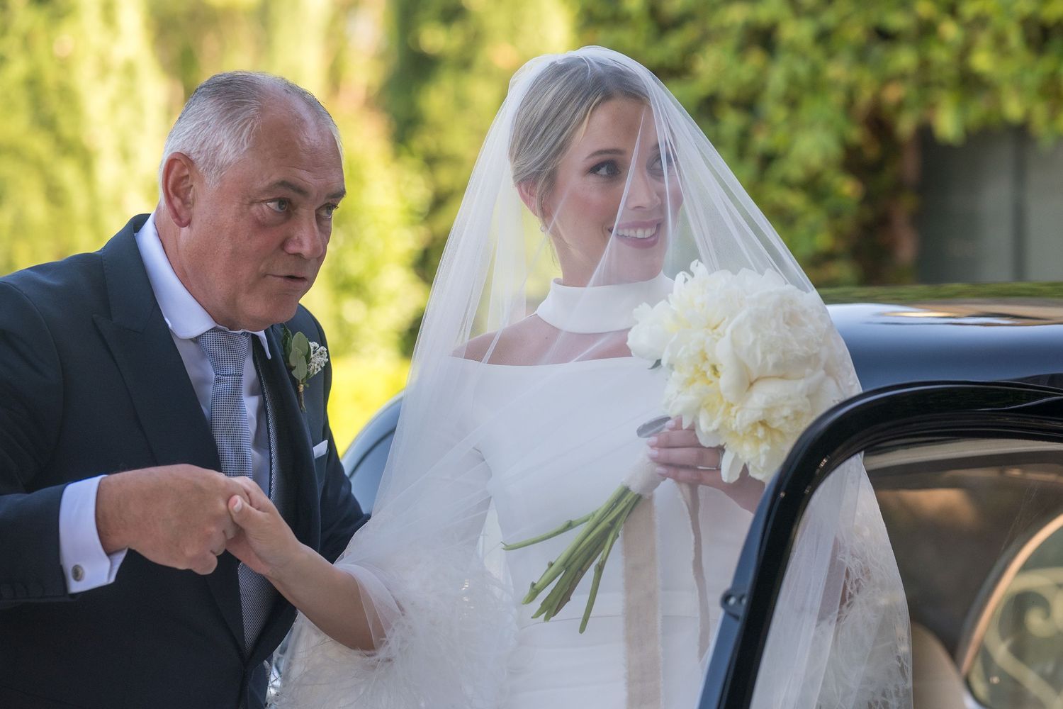 A person in a wedding dress holding white flowers exits a car accompanied by someone in formal attire.