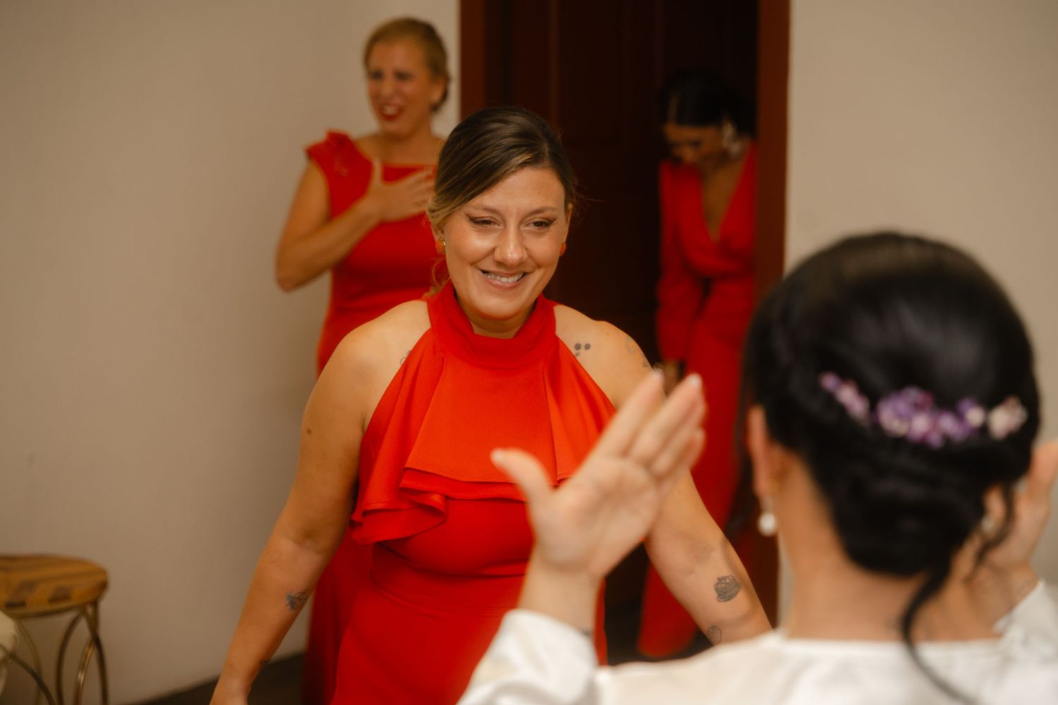 Two people in bright red dresses interact at an indoor event, with a third person visible in the background.