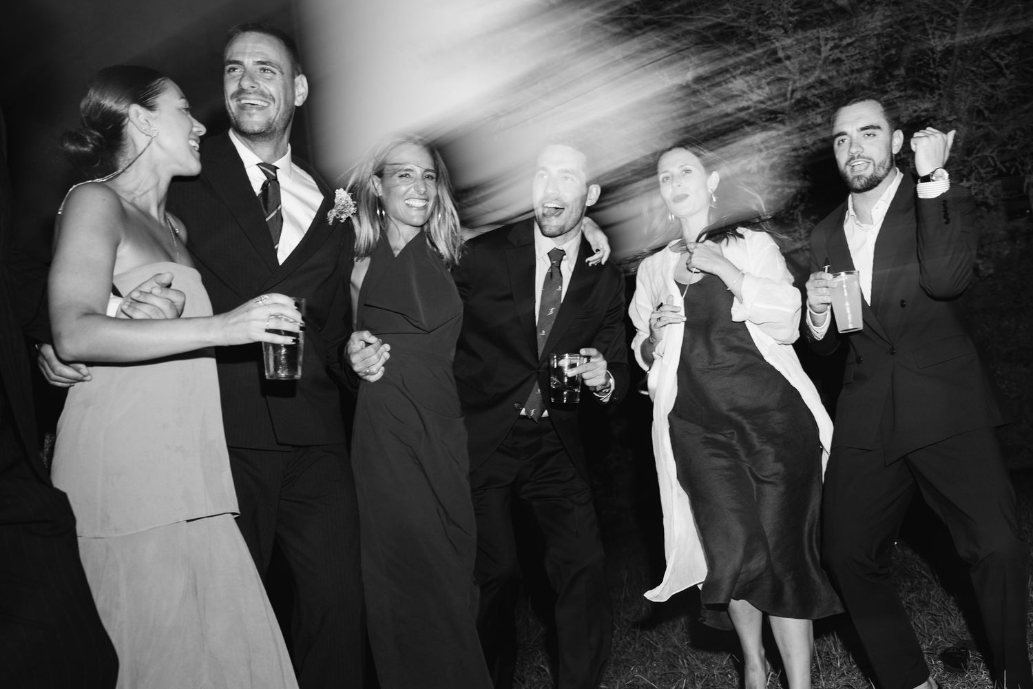 Black and white photo of a lively wedding reception with guests dancing and laughing together on the dance floor.