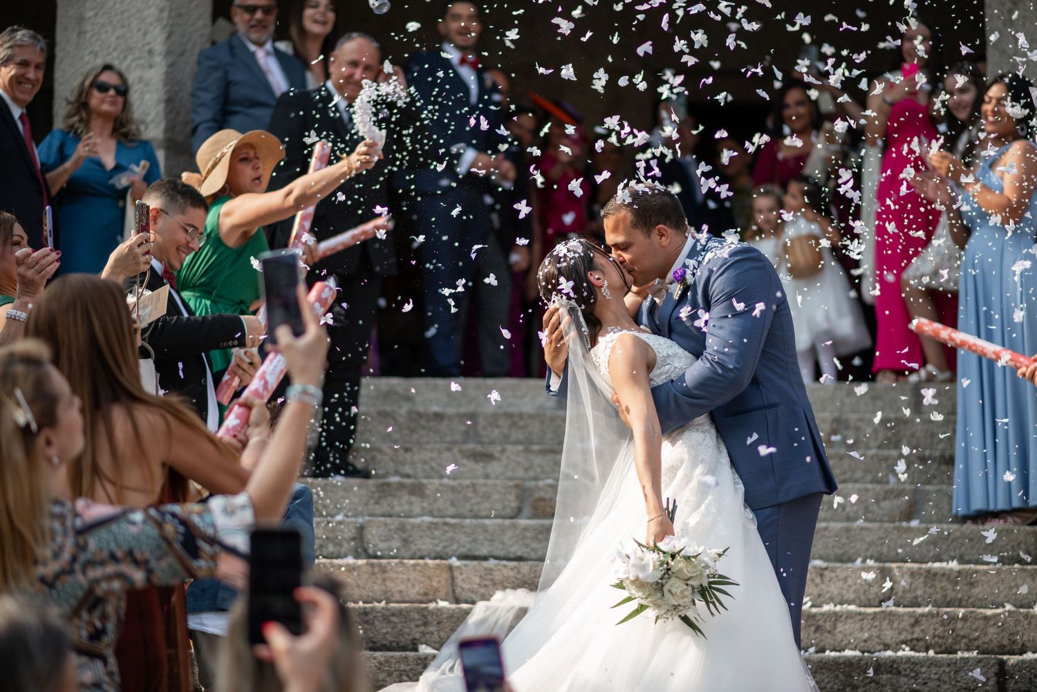 Wedding guests throw white confetti over a couple celebrating on stone steps during their special day.