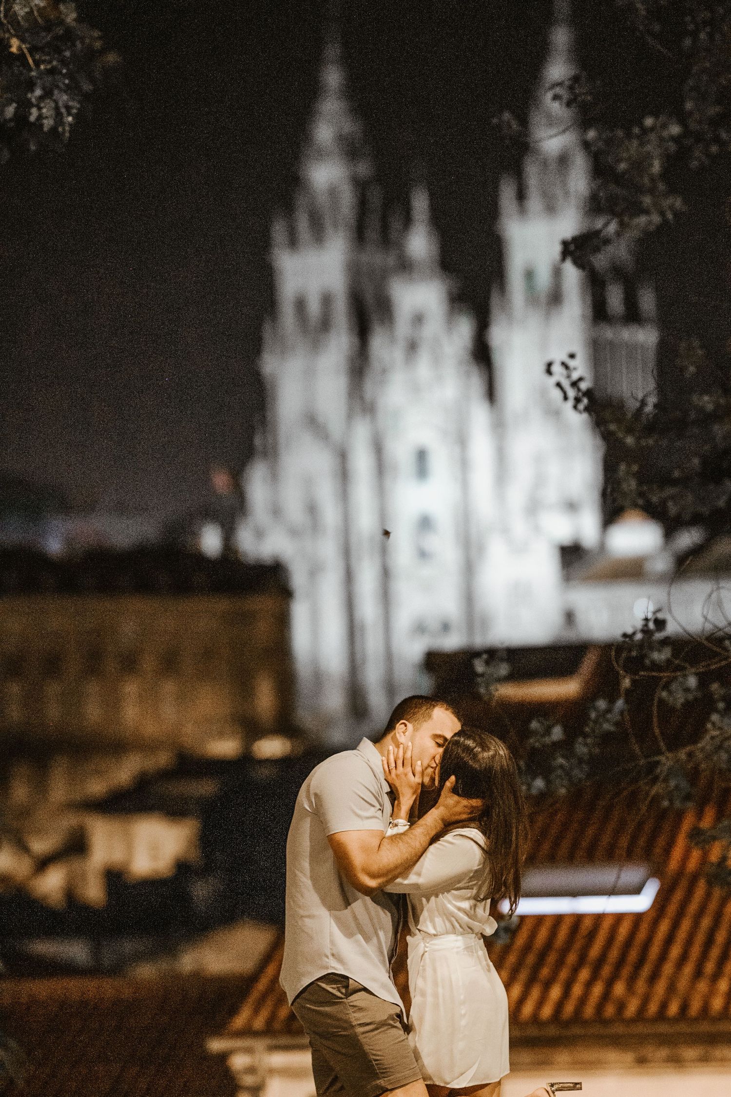 A romantic couple shares an intimate moment at night with an illuminated cathedral in the background.