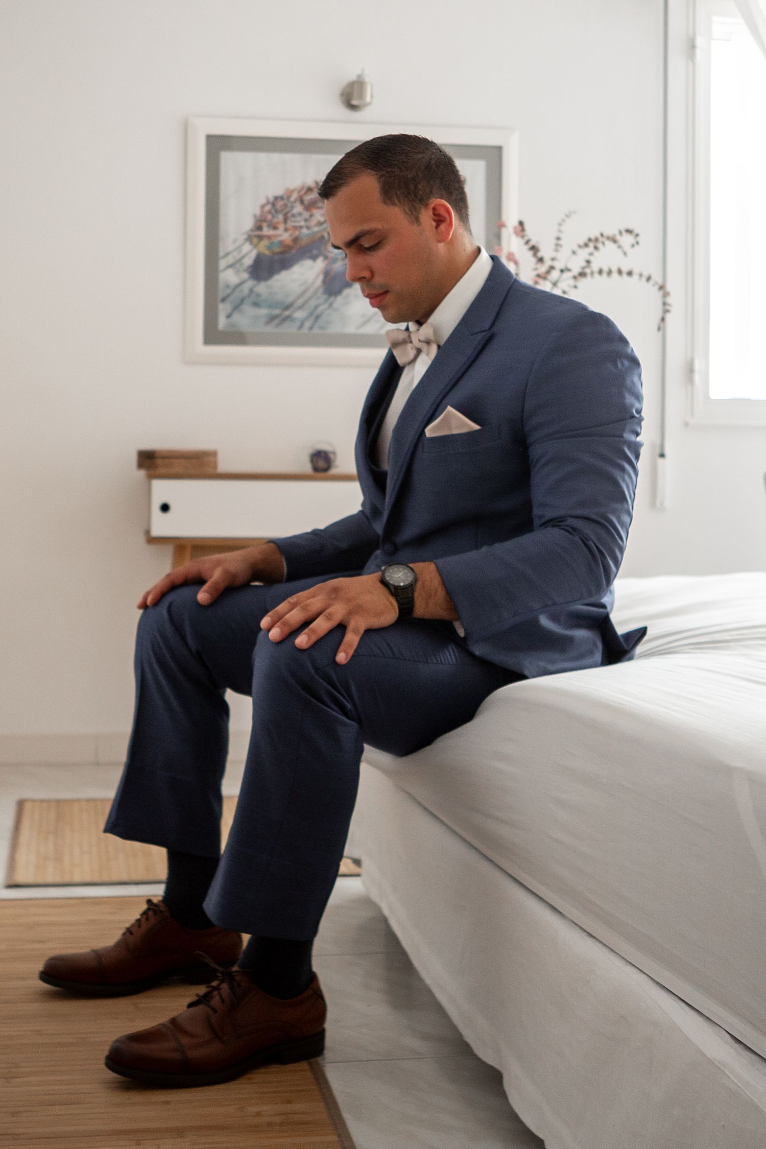 A person in a navy suit sits on a white bed, wearing brown boots in a minimalist bedroom setting.