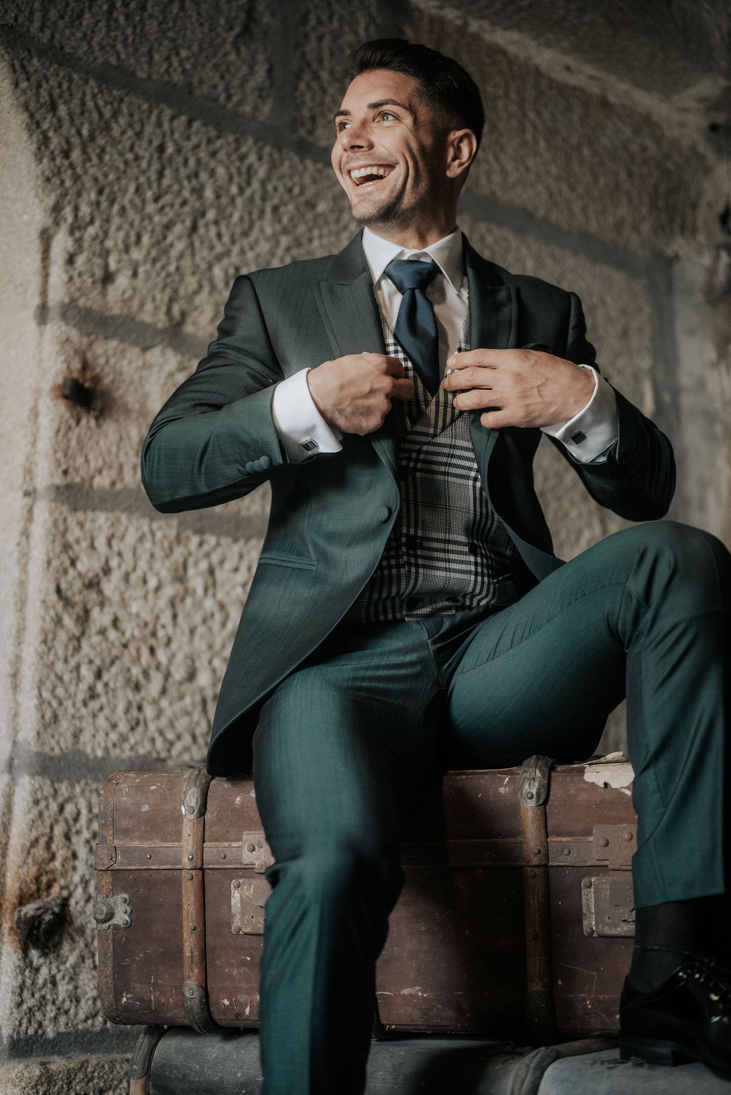 A person in a dark green suit and tie sits on a brick ledge while adjusting their vest and smiling.