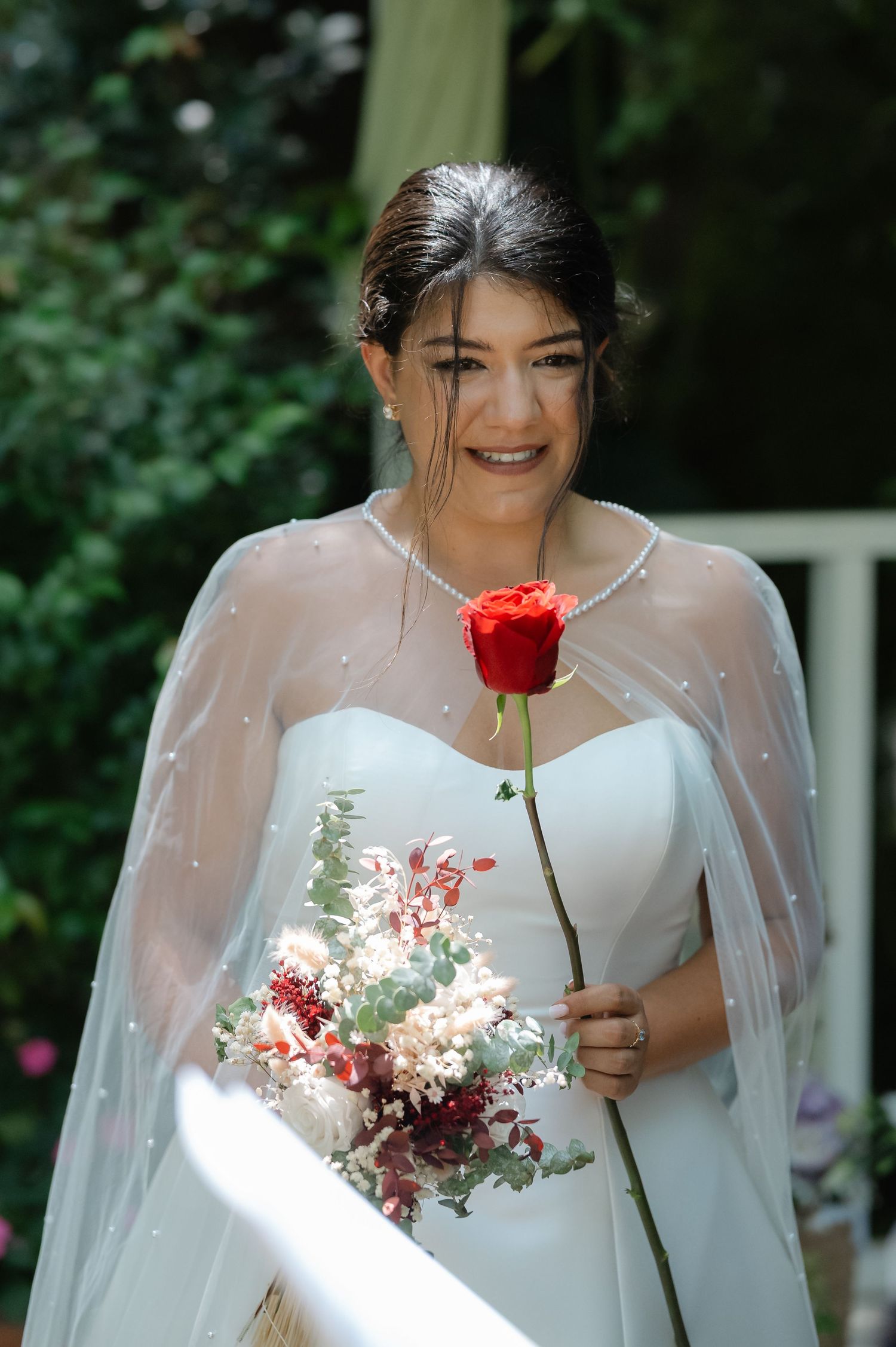 A bride in a white dress with sheer cape holds a single red rose against a garden backdrop.