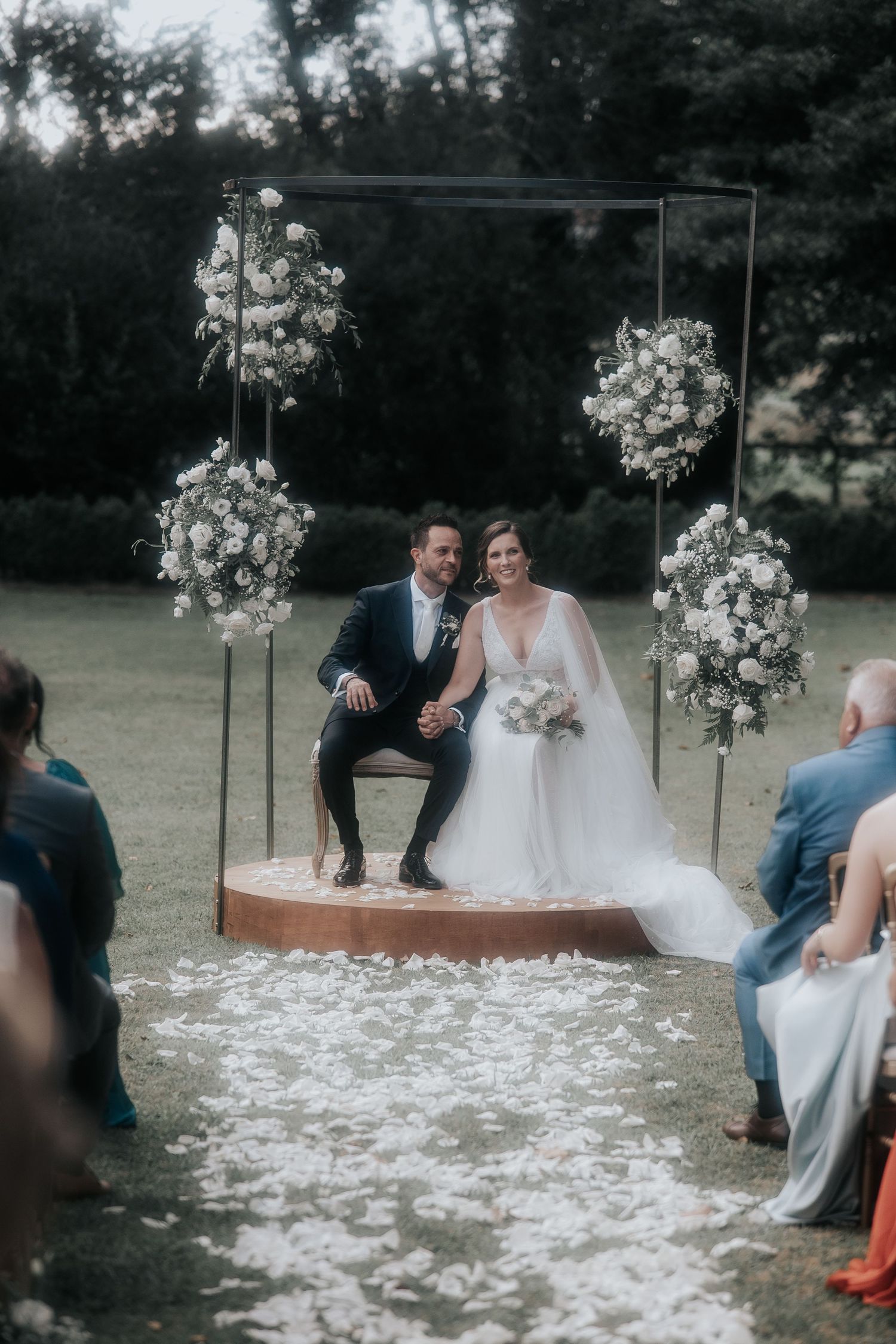Outdoor wedding ceremony setup with white floral arrangements and scattered petals on the ground.
