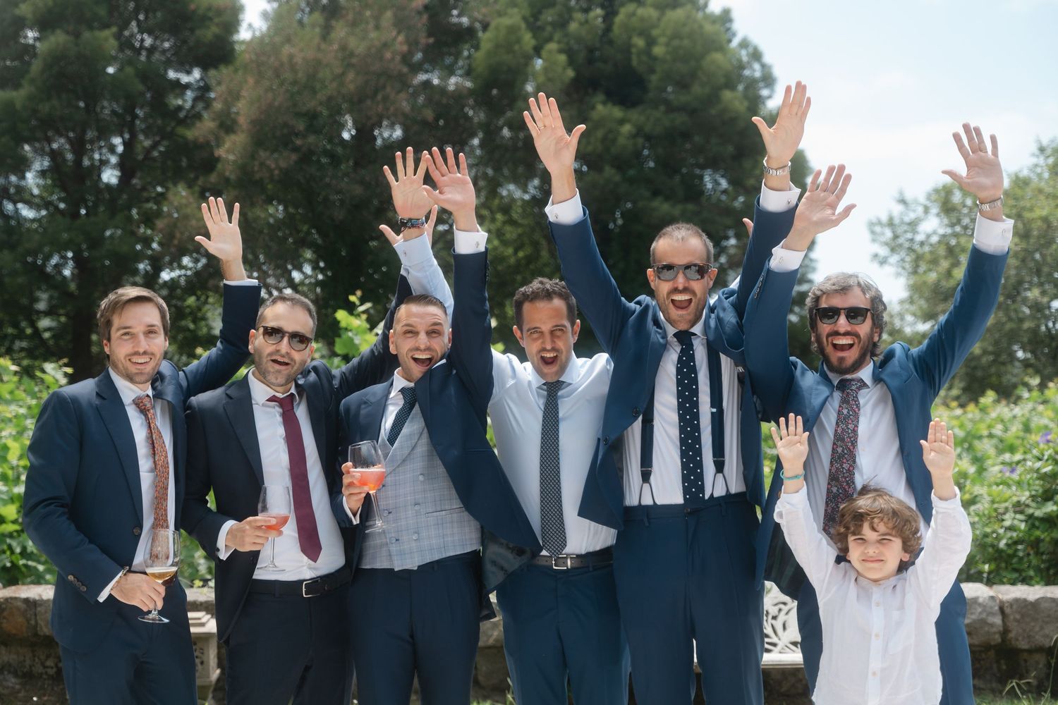 A group of formally dressed people raise their hands in celebration at an outdoor wedding event.