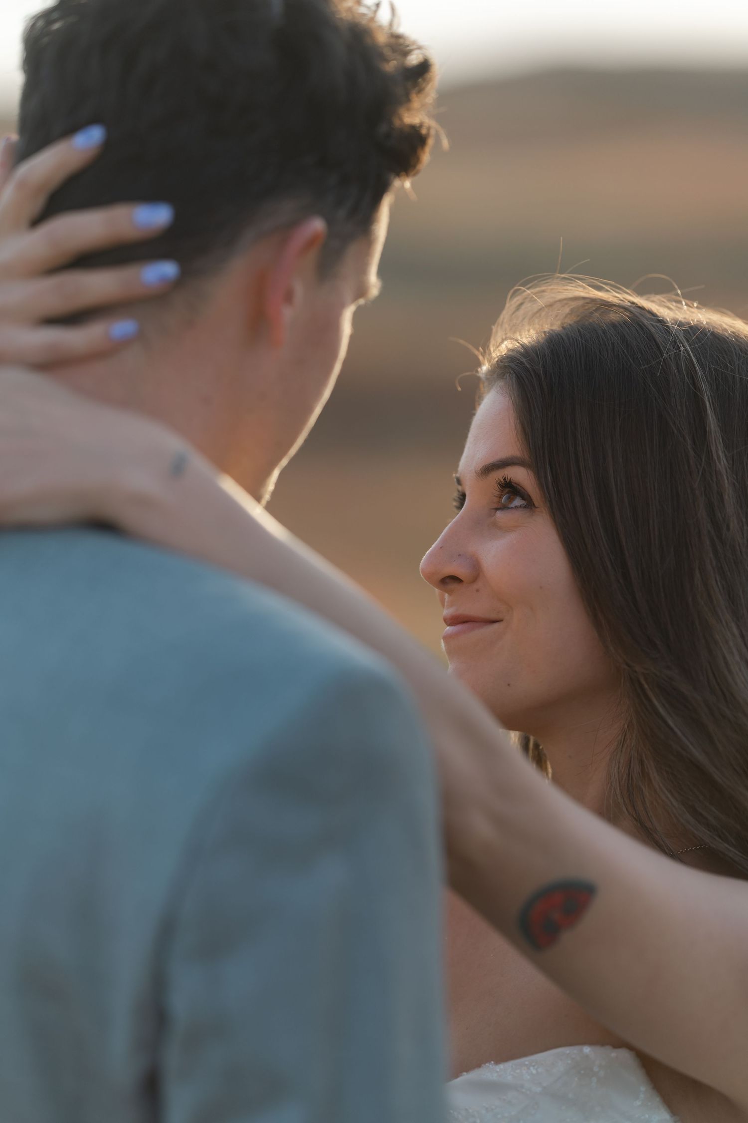 A smiling bride gazes lovingly at her groom while embracing him outdoors at sunset.