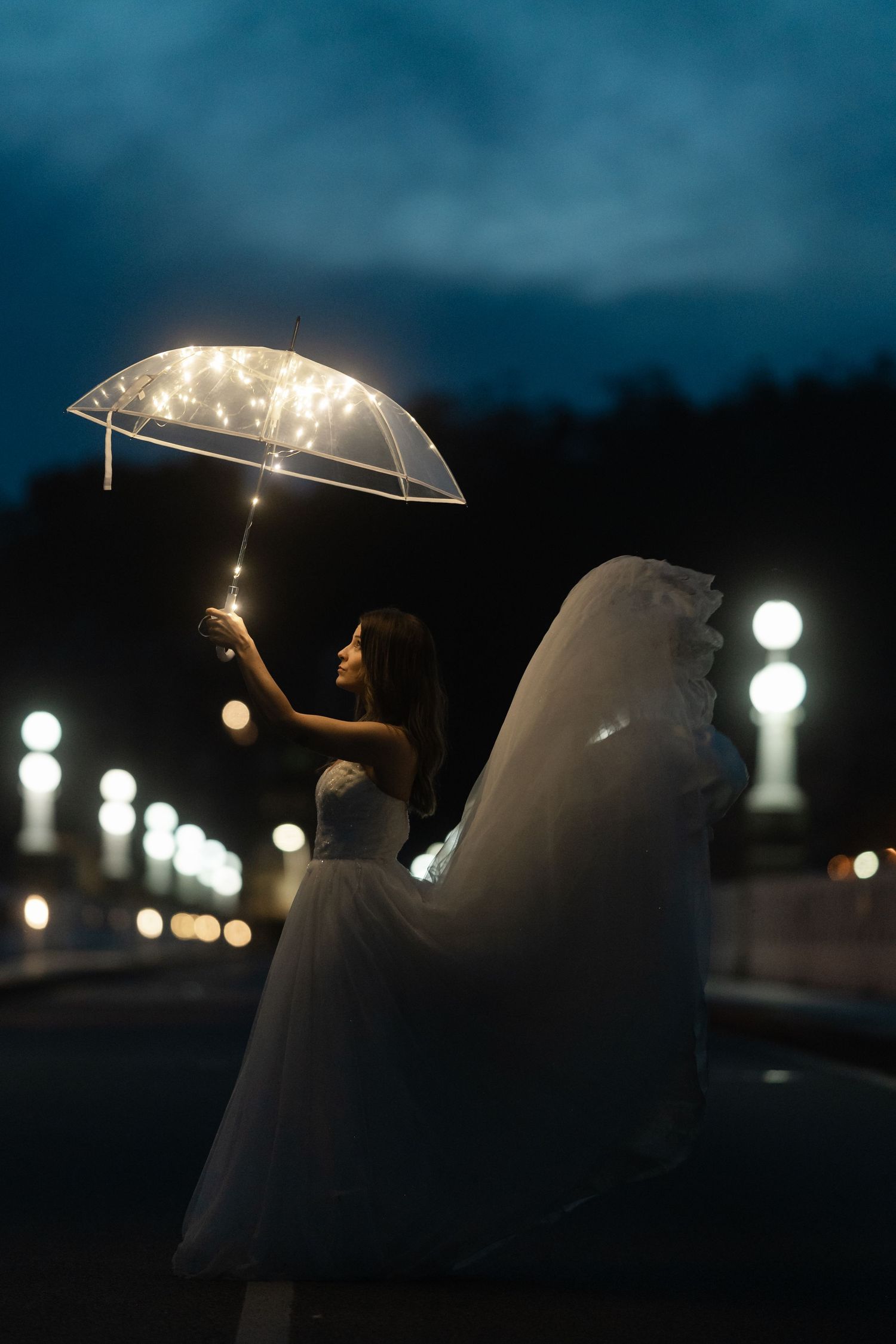 Bride in white gown holding illuminated transparent umbrella with fairy lights on a night street.