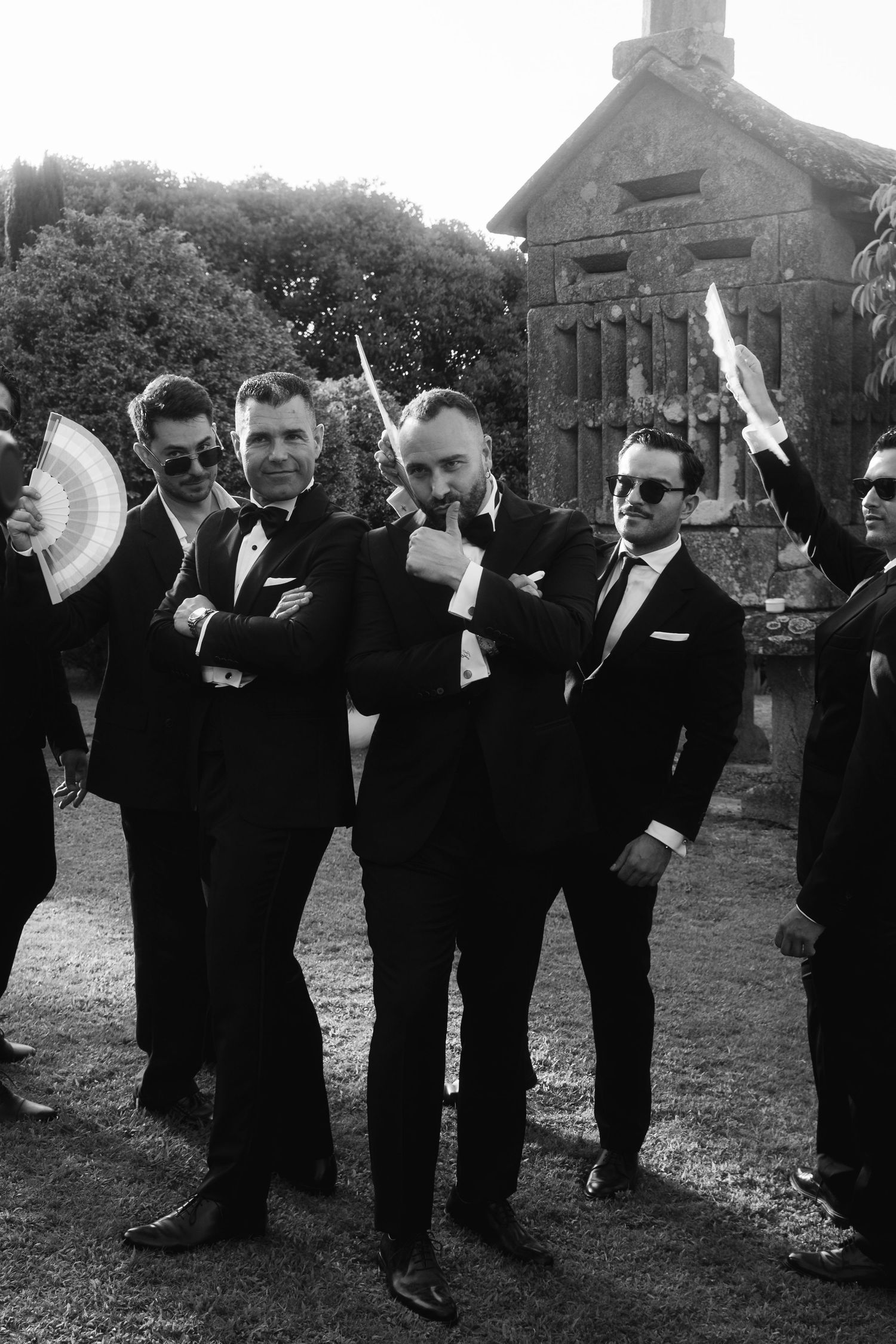 A group of groomsmen in black suits playfully pose together outdoors in a black and white photo.