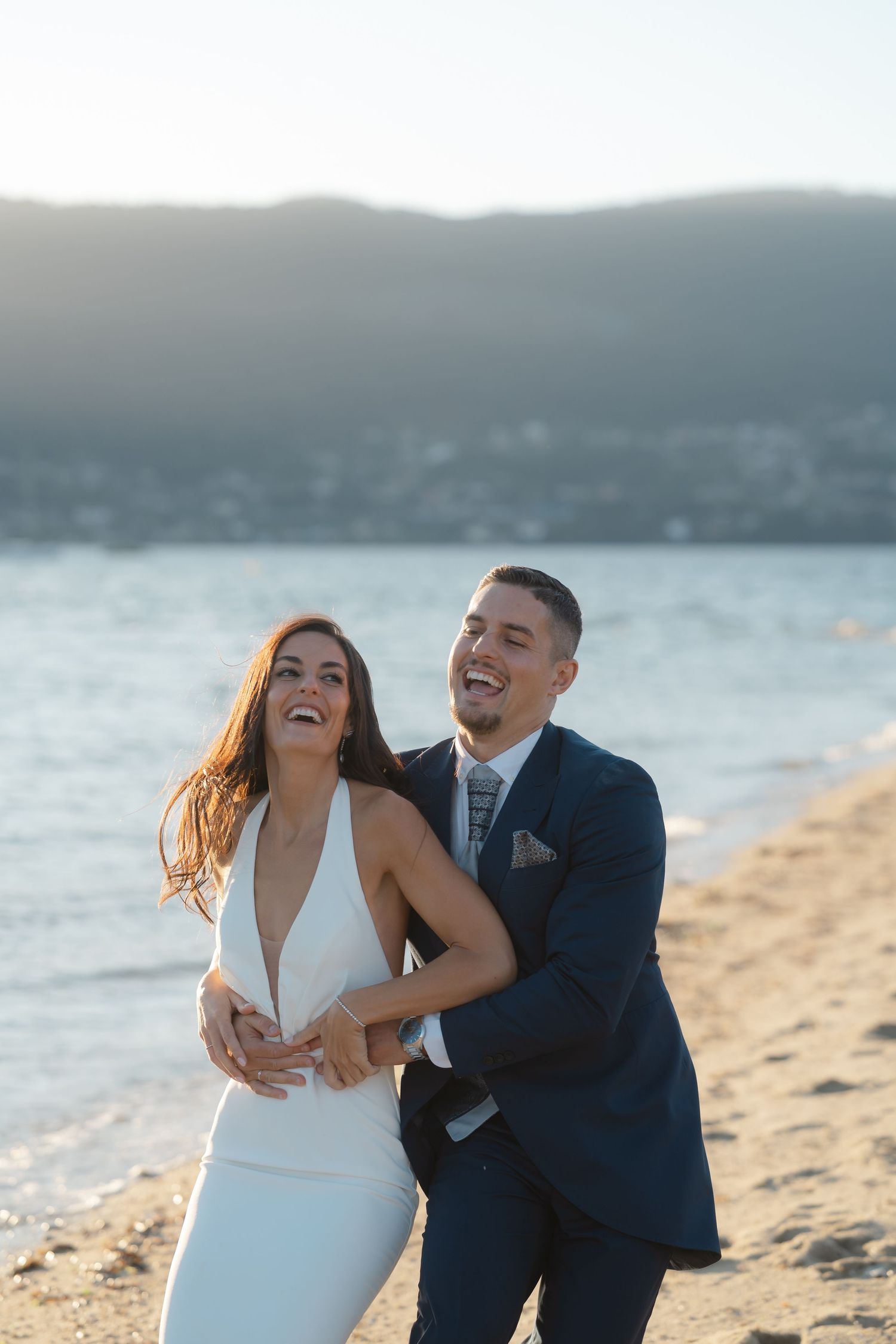 A happy couple shares joyful laughter together during their beach wedding photoshoot at sunset.