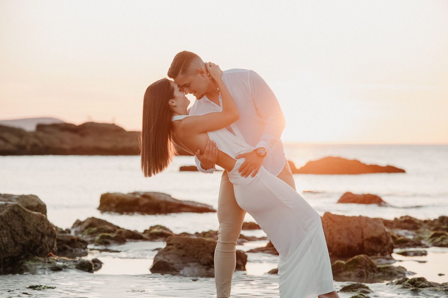 A romantic couple embraces on a rocky beach at sunset with ocean waves crashing in the background.