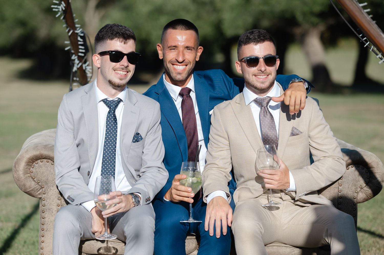 Three stylishly dressed friends in suits relax on outdoor furniture during a celebration.