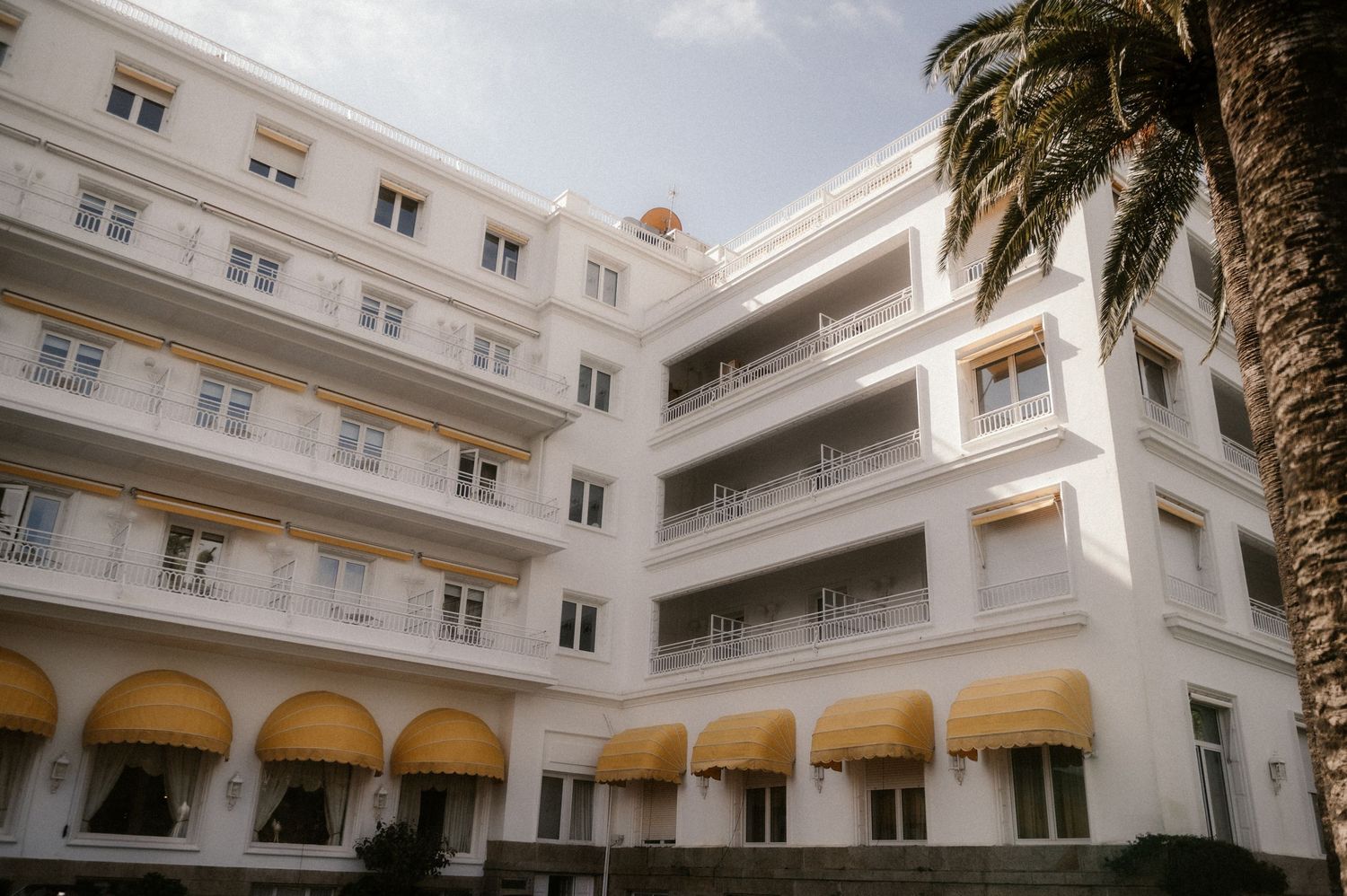 White colonial-style building with arched windows and balconies surrounds an interior courtyard with palm trees.