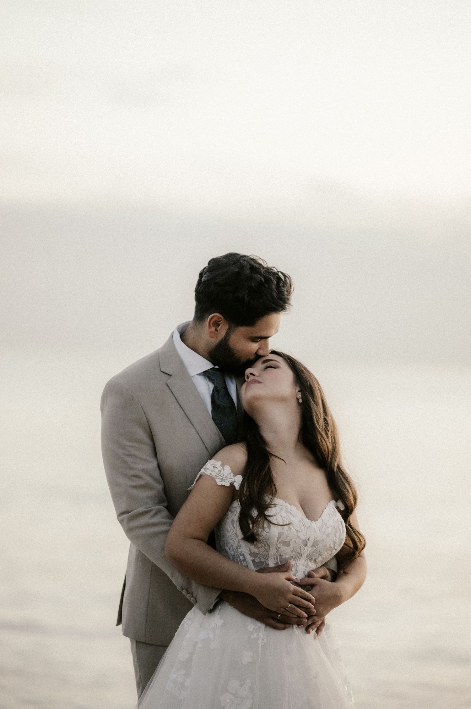 A romantic couple in wedding attire share tender kisses against a soft, misty backdrop in three similar poses.