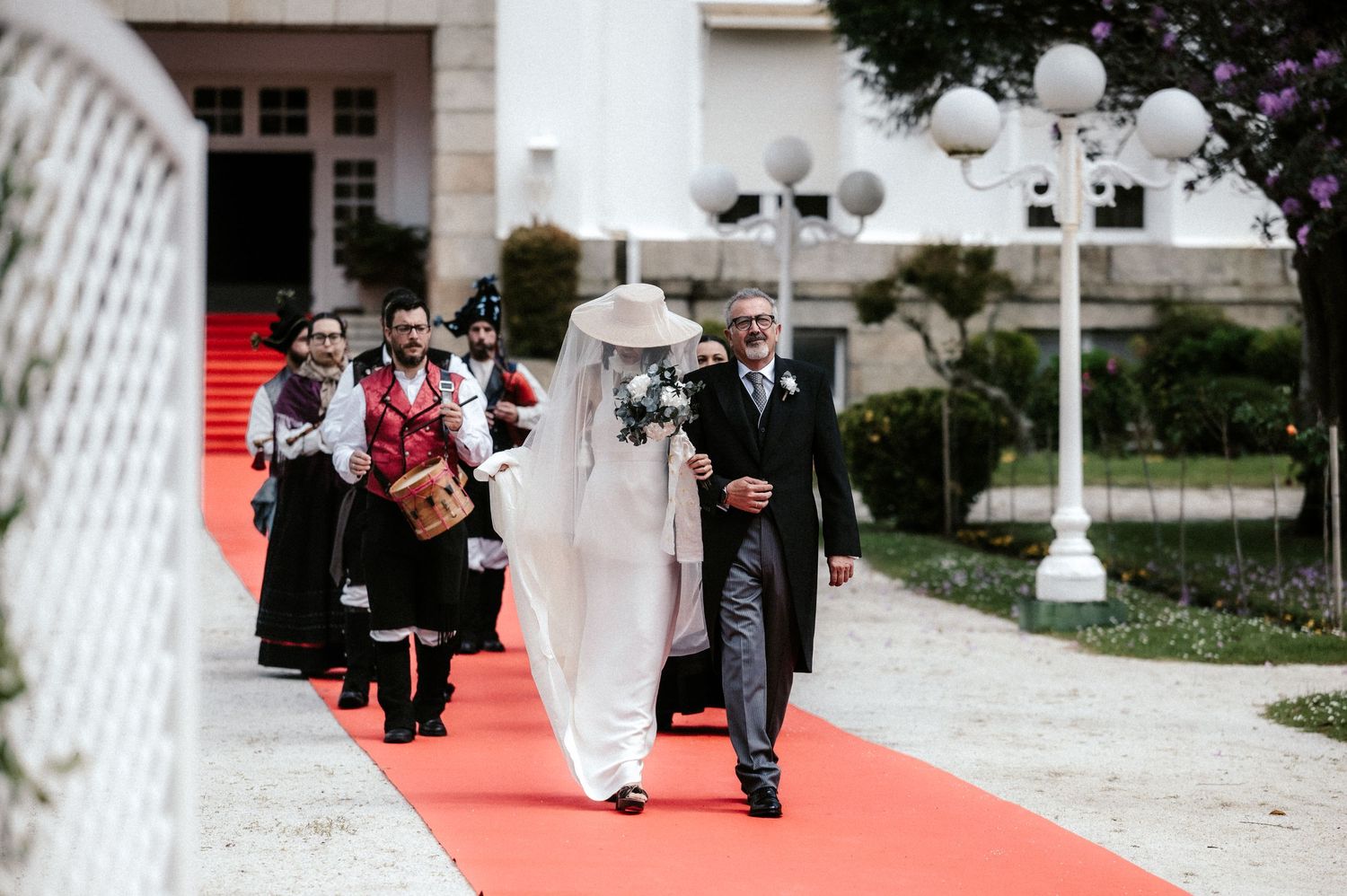 A couple walks down a red carpet pathway outside a white building with ornate lamp posts.