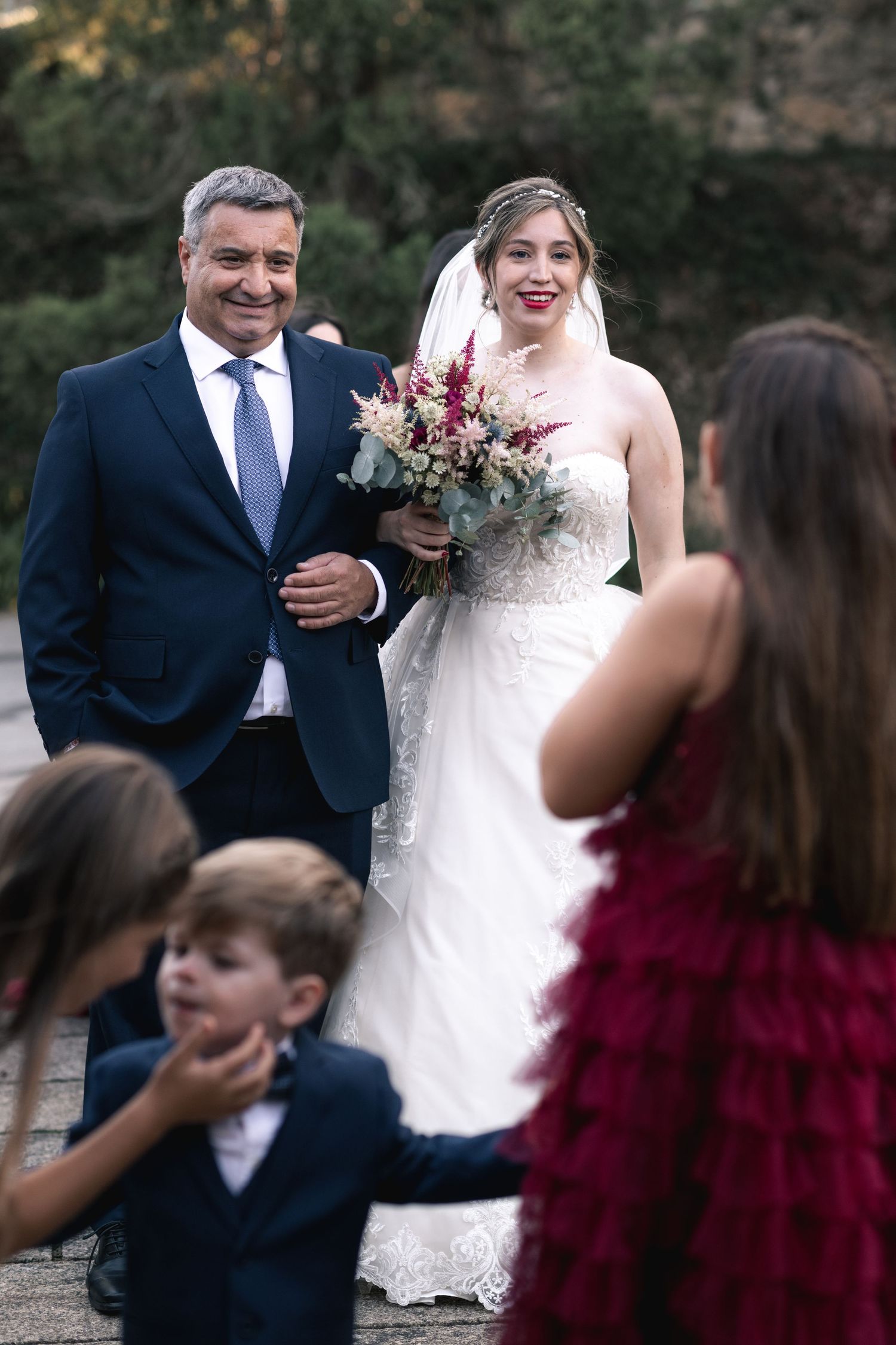 A wedding couple shares a candid moment while posing for photos with guests in burgundy attire outdoors.
