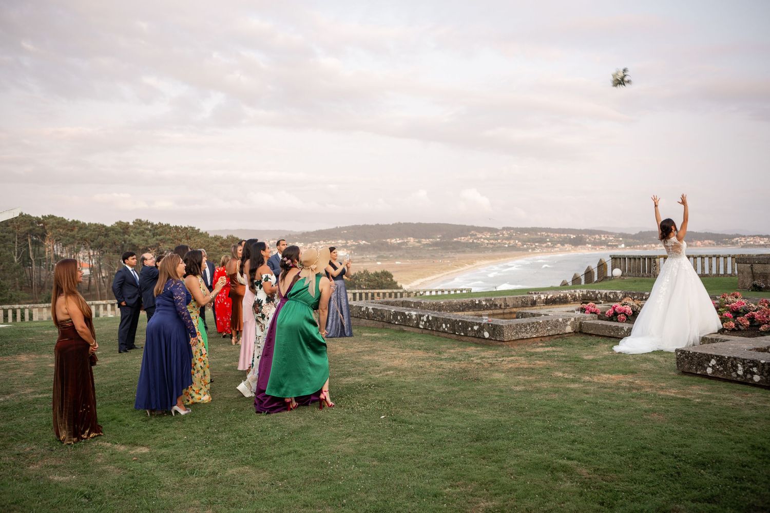 Wedding guests in formal attire gather on a hilltop lawn overlooking a scenic coastal view during an outdoor celebration.