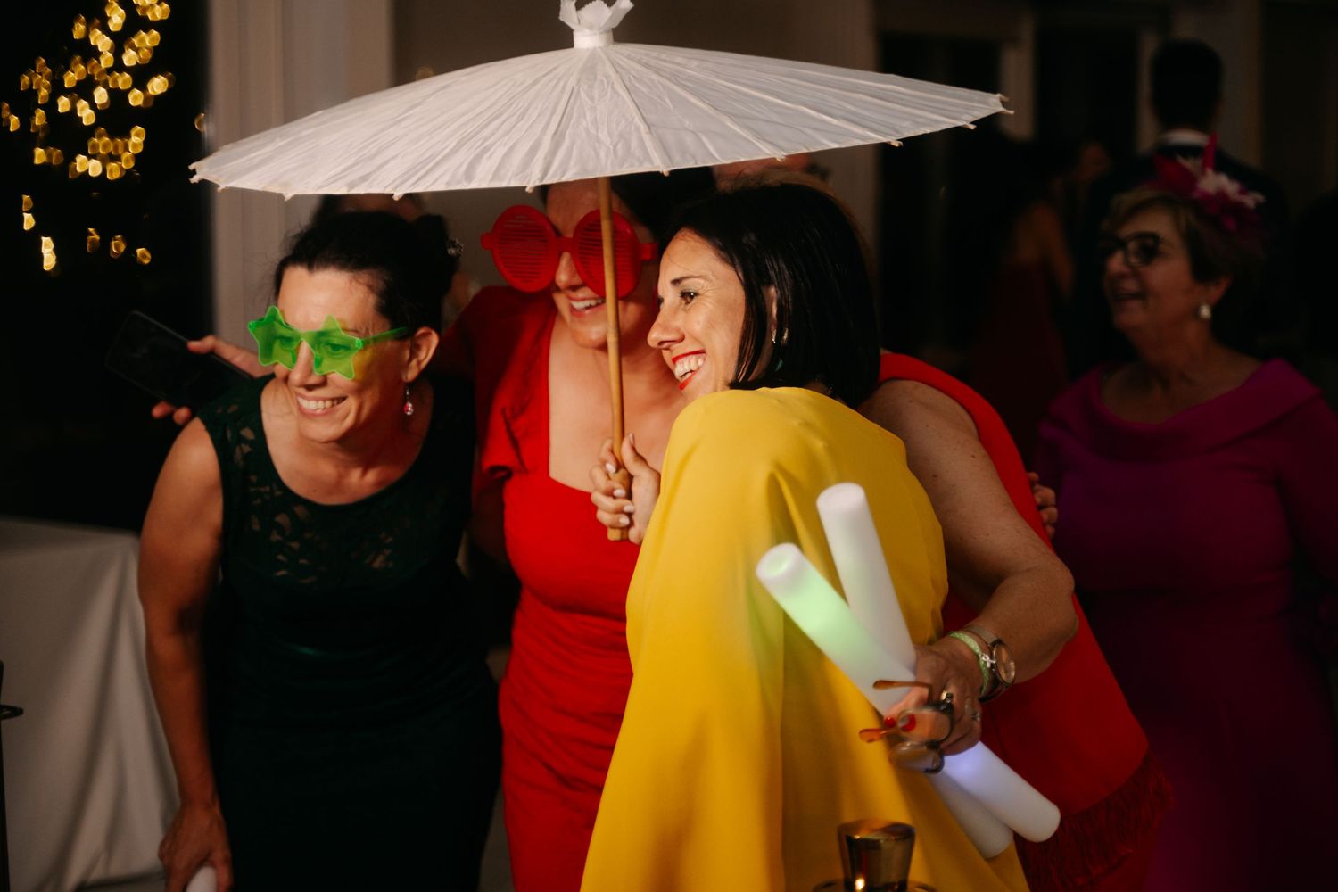 Wedding guests celebrate with party props including a white paper umbrella and neon green sunglasses.