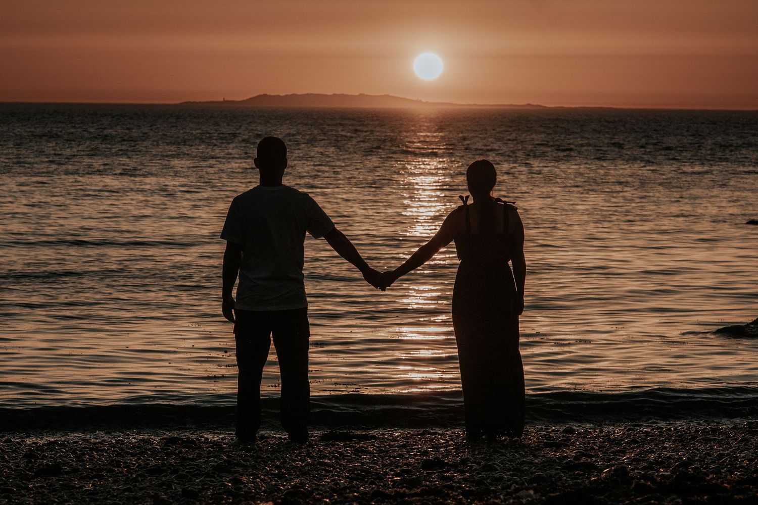 Silhouettes of people sitting by ocean at sunset with mountains in background.