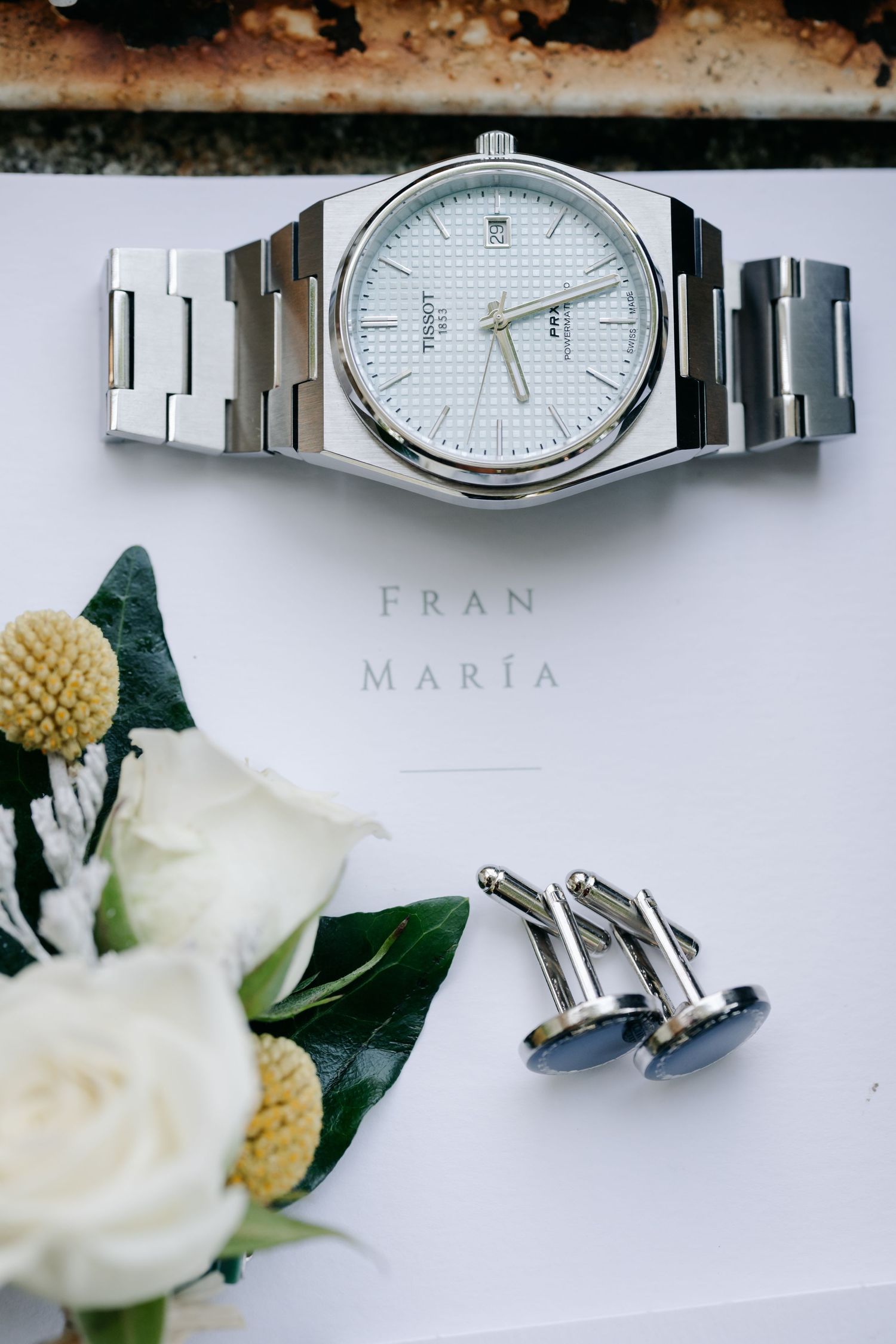 Silver analog watch with metal bracelet displayed alongside white flowers and blue cufflinks on a light background.