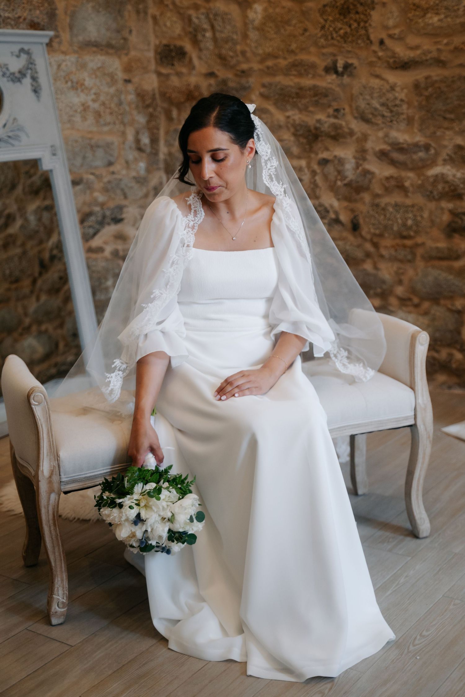 A bride in a classic white wedding gown sits gracefully in an elegant chair against a stone wall.