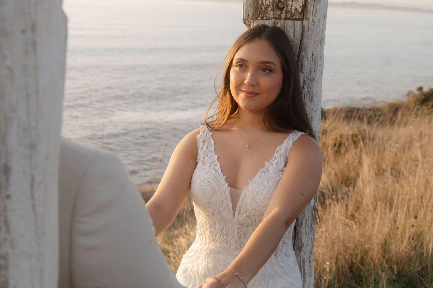 Bride in lace gown poses near white fence posts at seaside during sunset.
