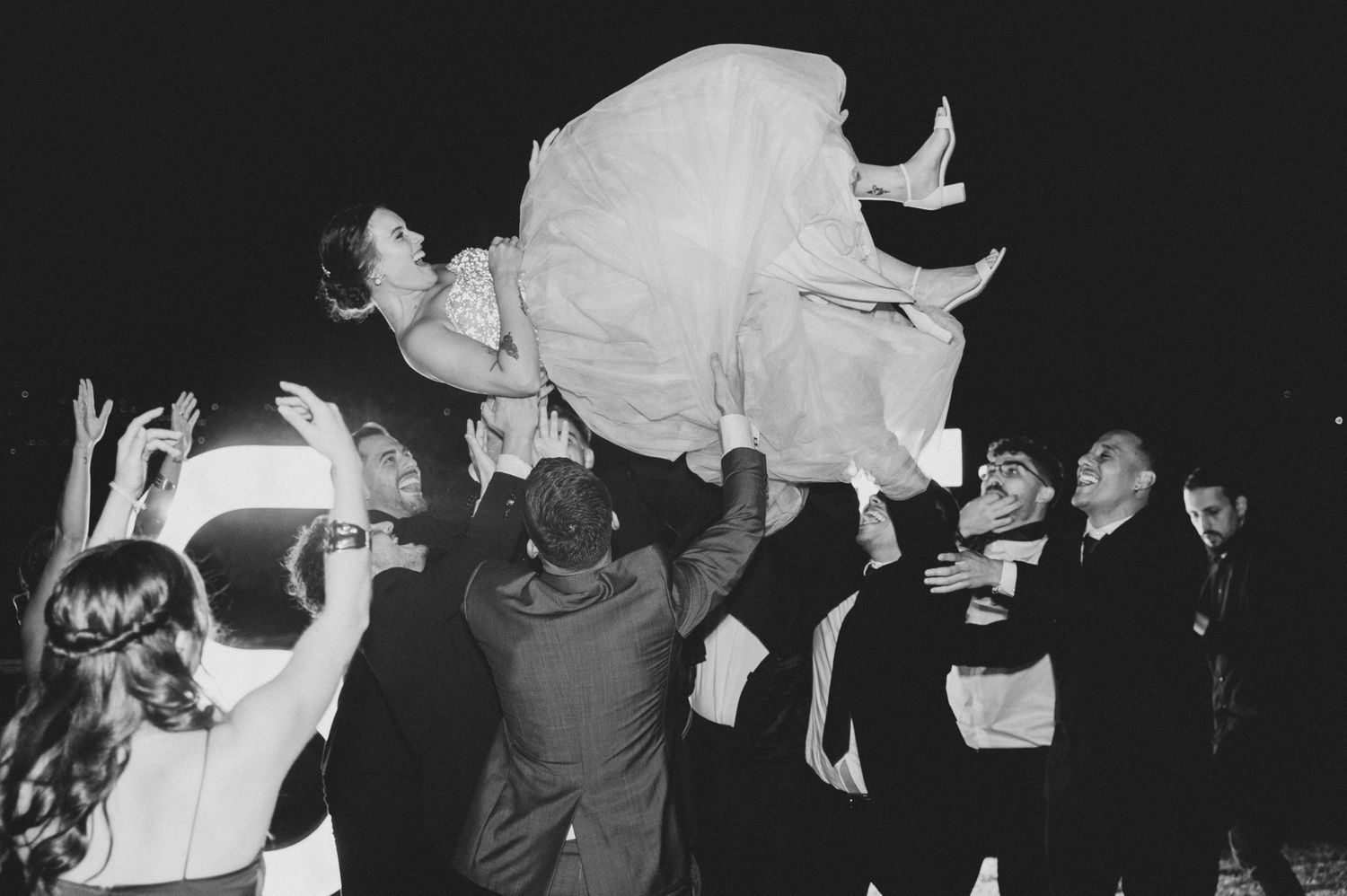 Wedding guests lift a bride in her white dress during a celebratory nighttime reception moment.