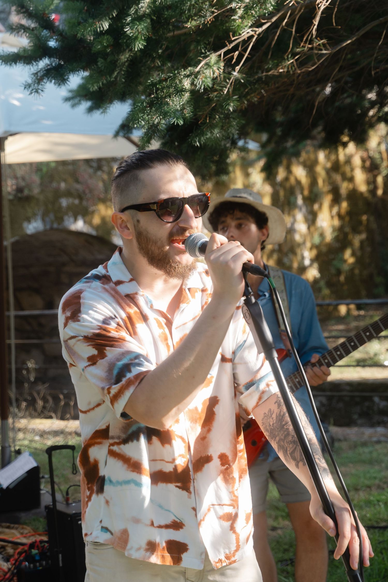 Singer in sunglasses and tie-dye shirt performs at an outdoor venue with guitarist in background.