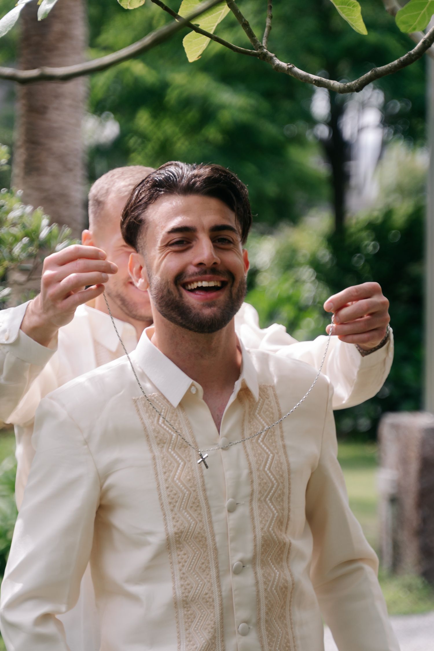 A person in a cream-colored traditional vest and shirt poses joyfully with raised arms in an outdoor setting.