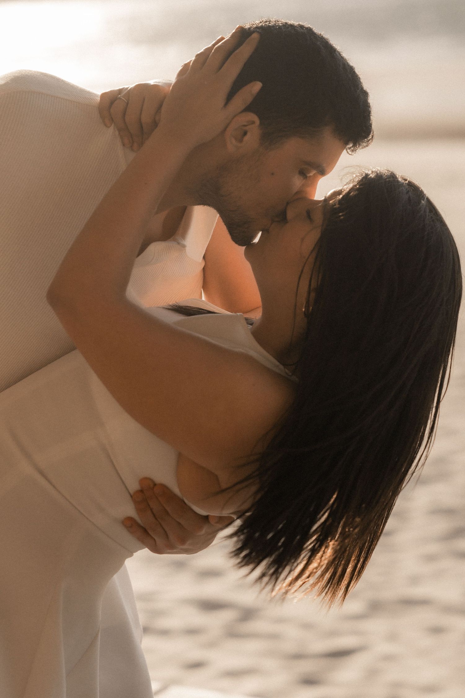 Intimate couple in white clothing share tender moments on a sunlit beach.
