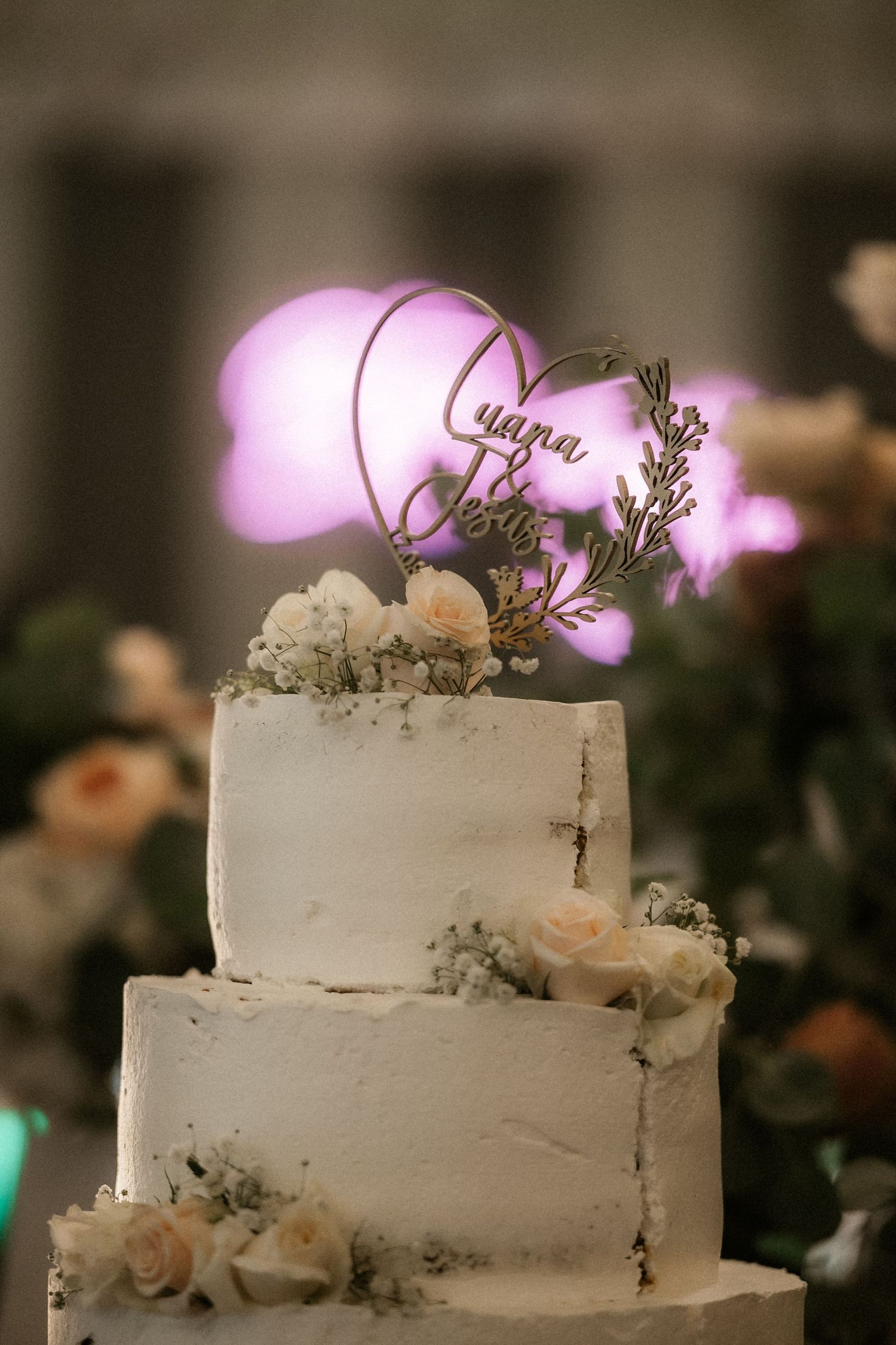 Three-tiered white wedding cake decorated with delicate roses and floral accents under purple ambient lighting.