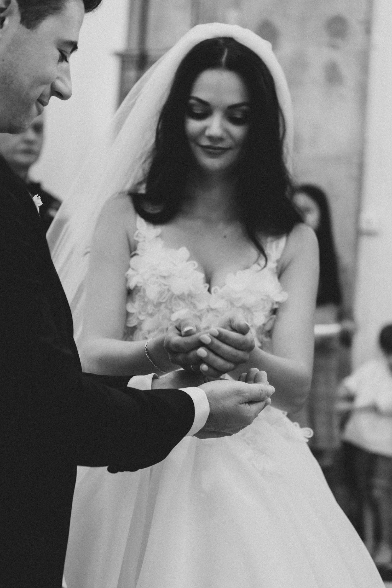 A bride in a white wedding dress with lace details smiles while exchanging rings during the ceremony.