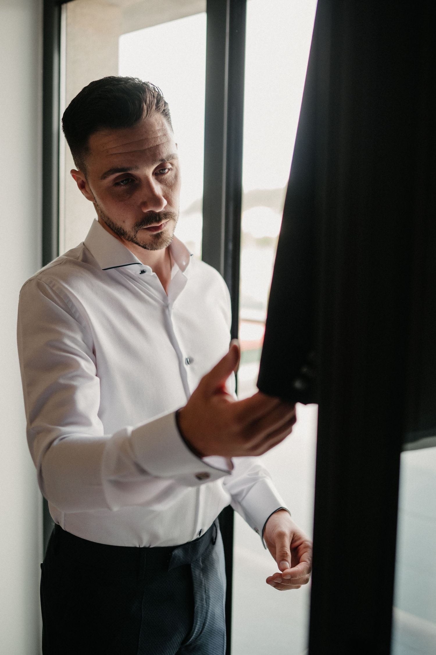 A person in a white dress shirt and dark pants gestures expressively while standing near a window.